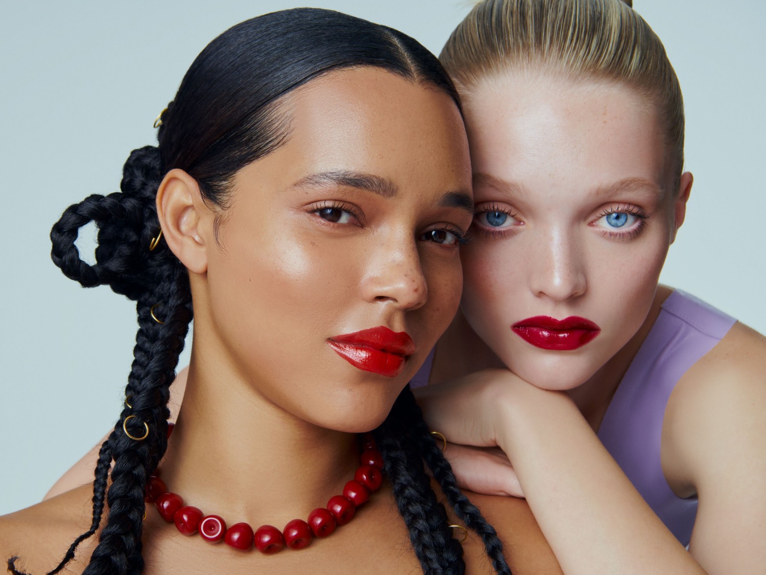 Nahaufnahme von zwei Models mit glänzenden roten Lippen, wobei das eine dunkle Zöpfe und eine rote Halskette trägt und das andere hellblonde Haare und blaue Augen hat.