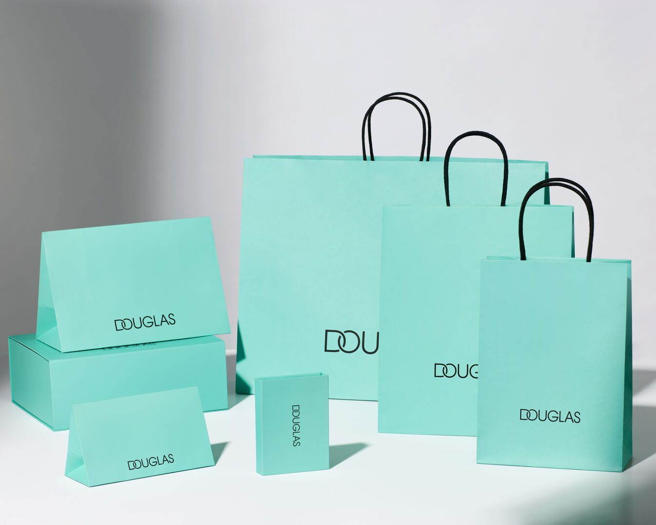 Na zdjęciu widoczne są torebki i pudełka Douglas w kolorze turkusowym, z czarnym logo, tworzące elegancką kompozycję.