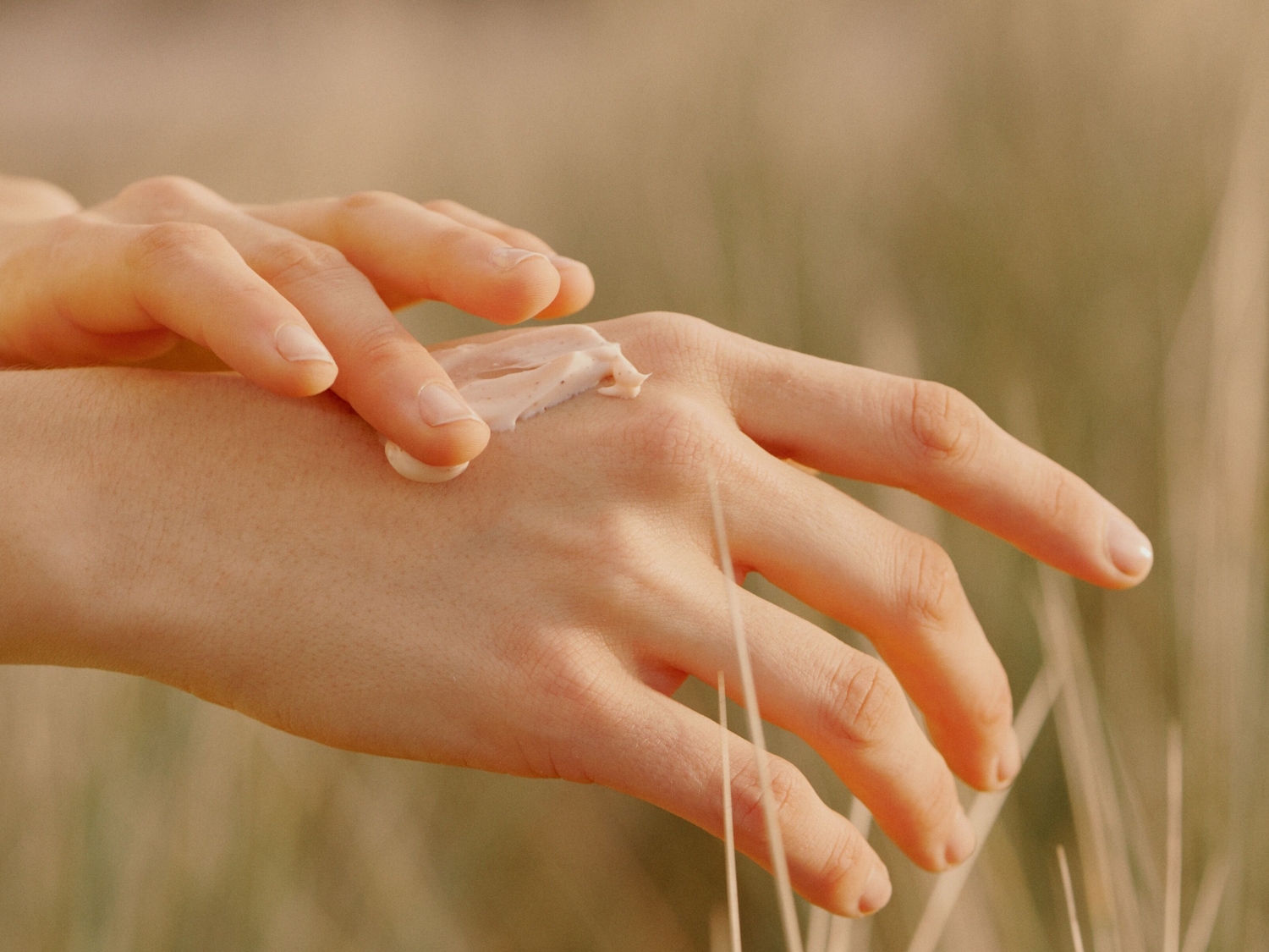 Nahaufnahme einer Hand, die eine beigefarbene Creme auf den Handrücken aufträgt, umgeben von Gräsern.