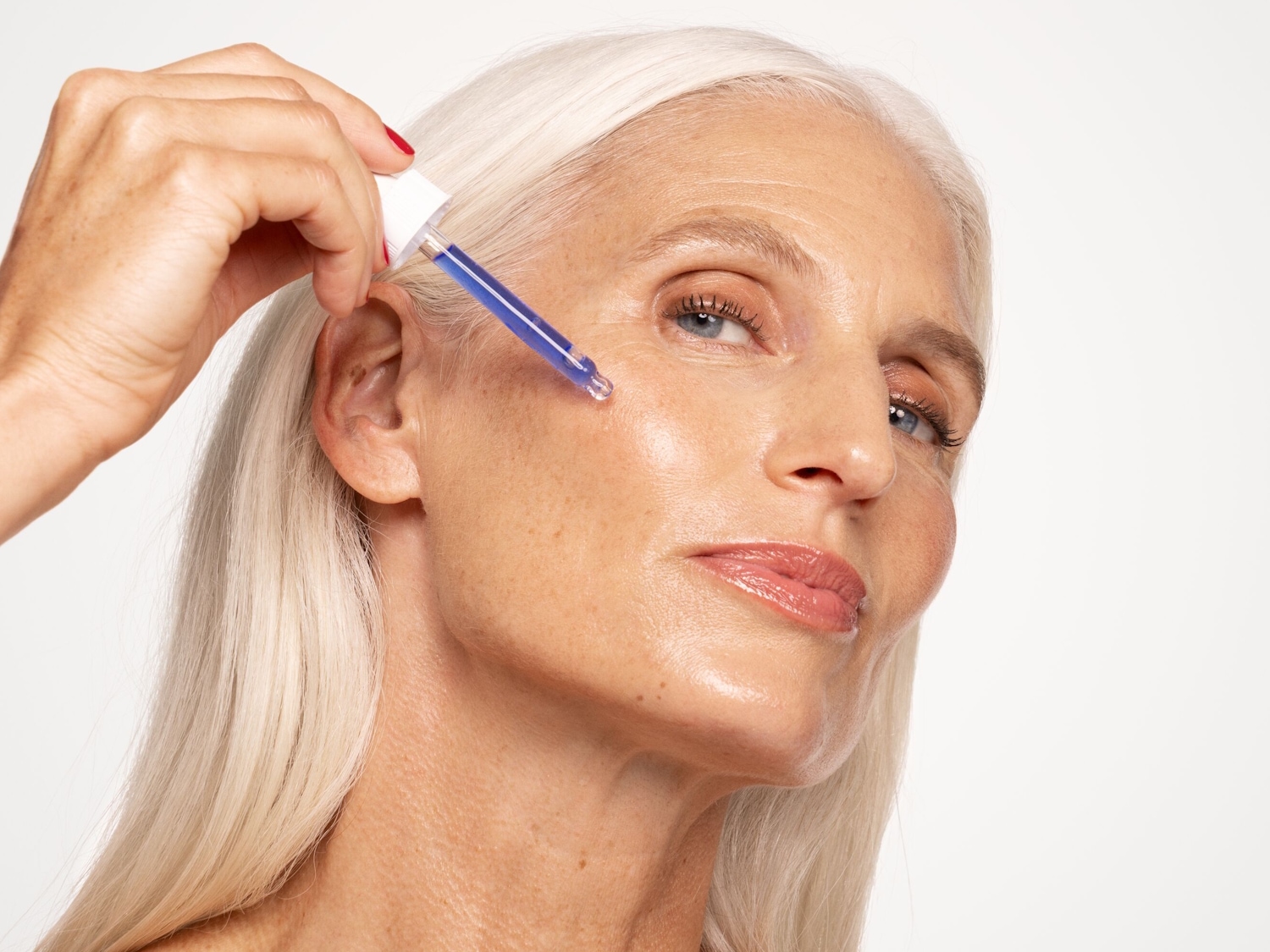 Eine Frau mit silberblondem Haar trägt ein Serum mit einer Pipette auf ihr Gesicht auf, um ihre Haut zu pflegen.