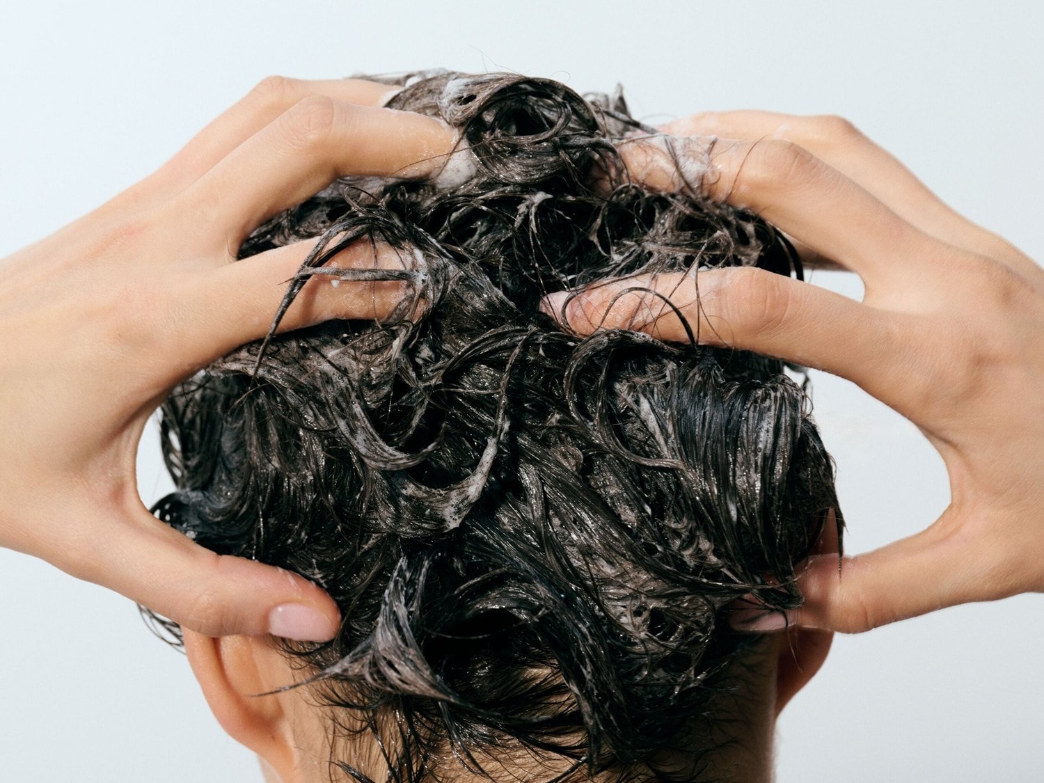 Zwei Hände massieren sanft schäumendes Shampoo in dunkles, nasses Haar, um es gründlich zu reinigen und zu pflegen.