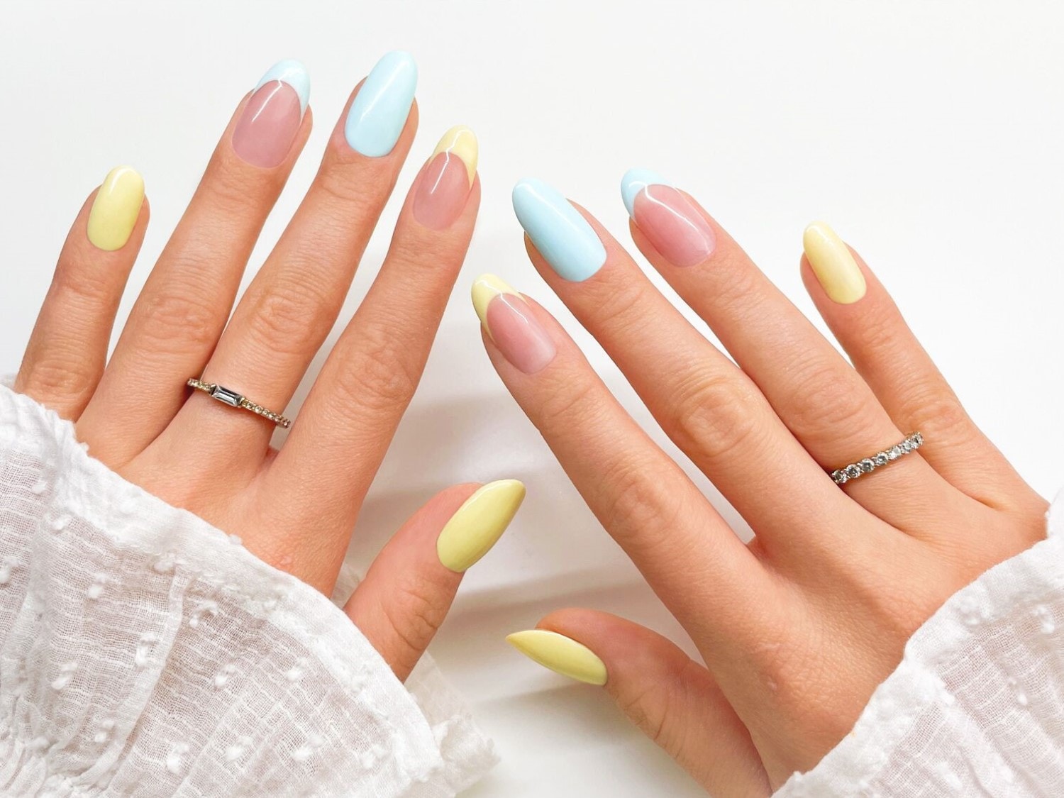 Zwei Hände mit pastellfarbenen Nägeln, die abwechselnd hellblau und gelb lackiert sind, wobei einige Nägel eine französische Spitze in diesen Farben aufweisen, und an jedem Ringfinger ist ein zarter Ring zu sehen.