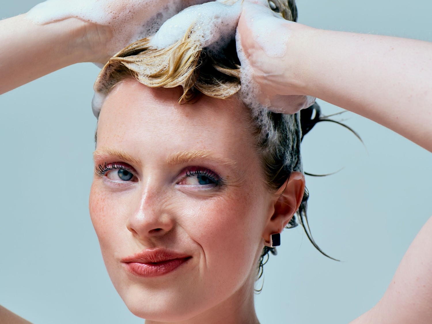 Eine Frau mit Sommersprossen massiert ihr blondes, schaumiges Haar unter der Dusche und blickt mit einem leichten Lächeln in die Kamera.