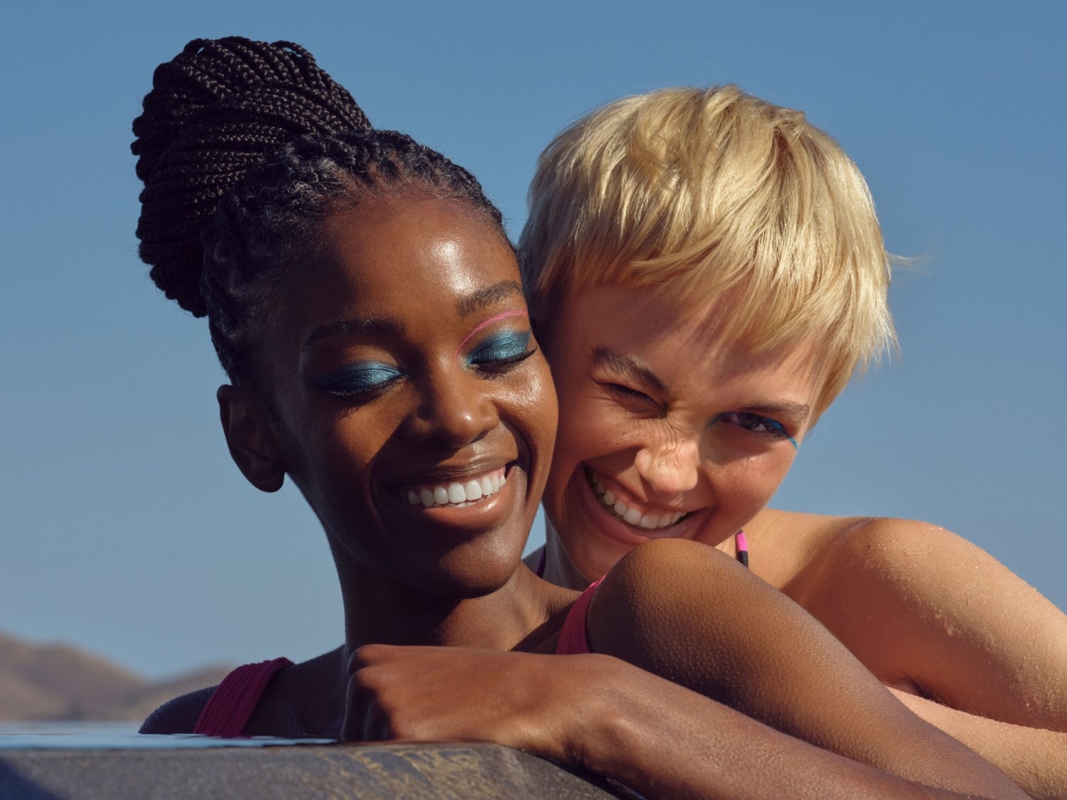 Zwei strahlende Frauen, die eine mit geflochtenem Haar und blauem Lidschatten, die andere mit blonden, kurzen Haaren und einem Augenzwinkern, genießen die Sonne.
