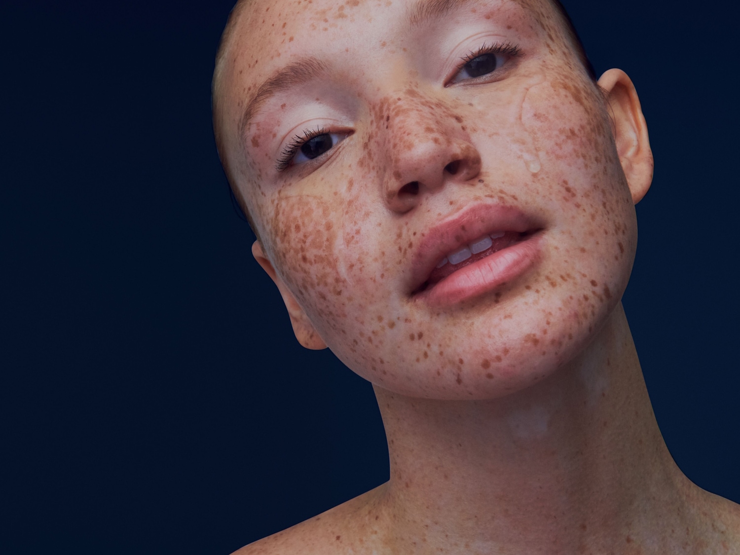 Nahaufnahme eines jungen, hellhäutigen Mädchens mit Sommersprossen und einem einzelnen Wassertropfen auf der Wange vor einem dunkelblauen Hintergrund.
