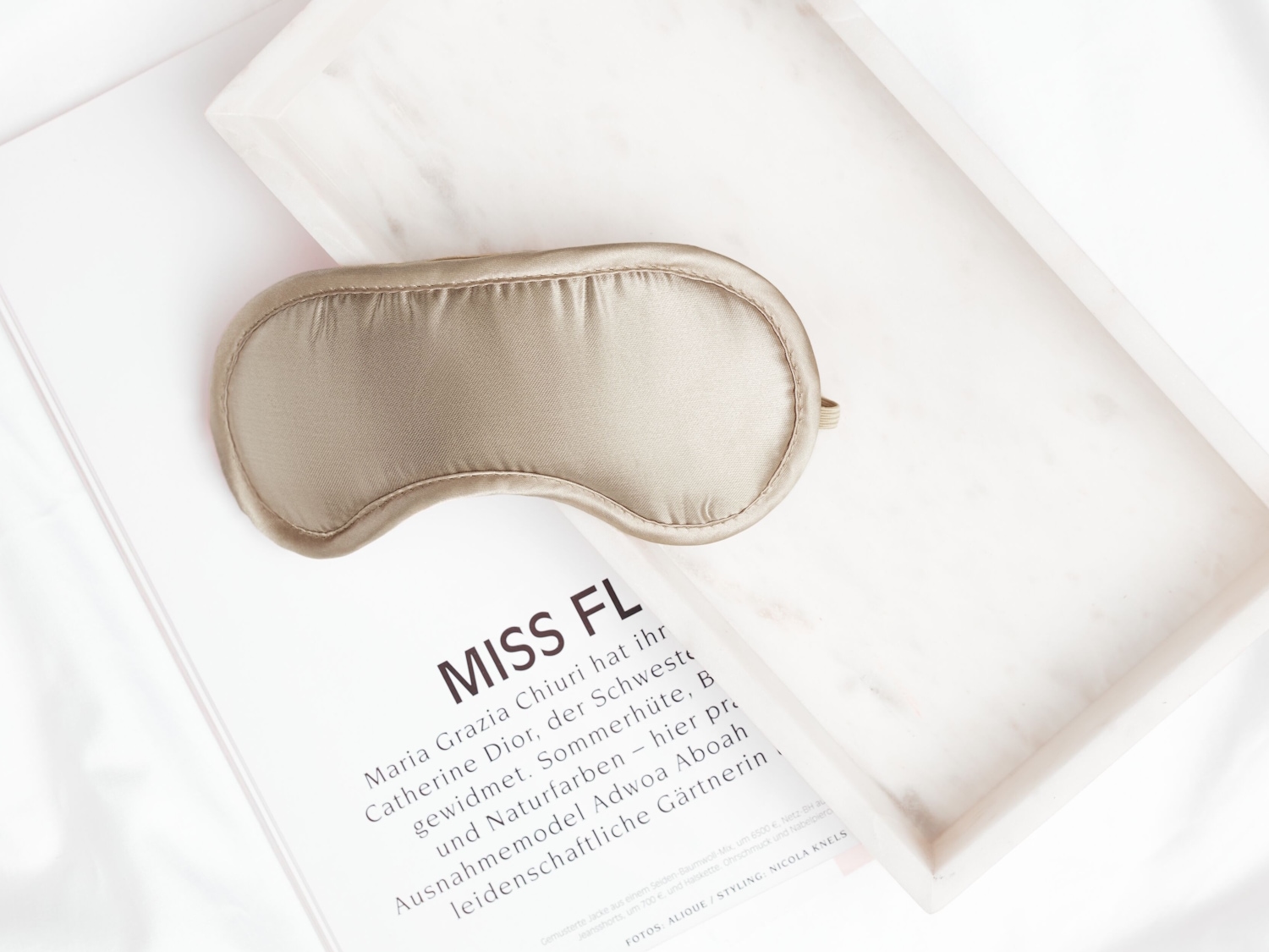 Eine goldene Schlafmaske liegt auf einer Zeitschrift und einem weißen Marmortablett.