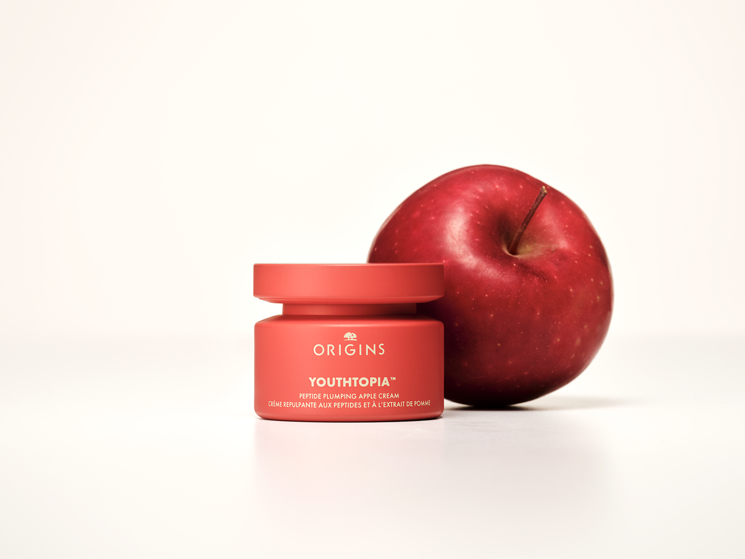 Een rode pot Origins Youthtopia peptide plumping appelcrème staat naast een rode appel, wat de natuurlijke ingrediënten en de jeugdherstellende eigenschappen van de crème benadrukt.