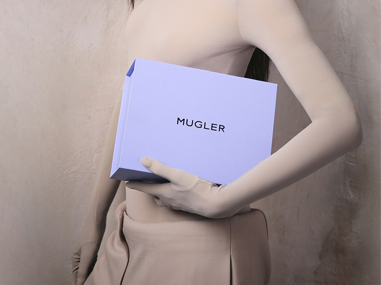 Een vrouw in een beige outfit met lange mouwen en handschoenen houdt een lichtpaarse doos met het zwarte woord "MUGLER" erop.