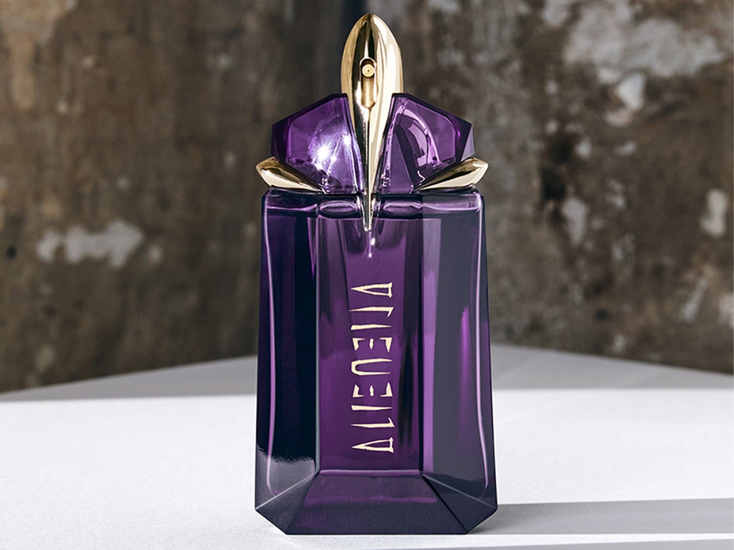Een paarse parfumfles met gouden accenten en de naam "ALIEN" verticaal in goudkleurige letters, staat op een witte ondergrond met een grijze achtergrond.