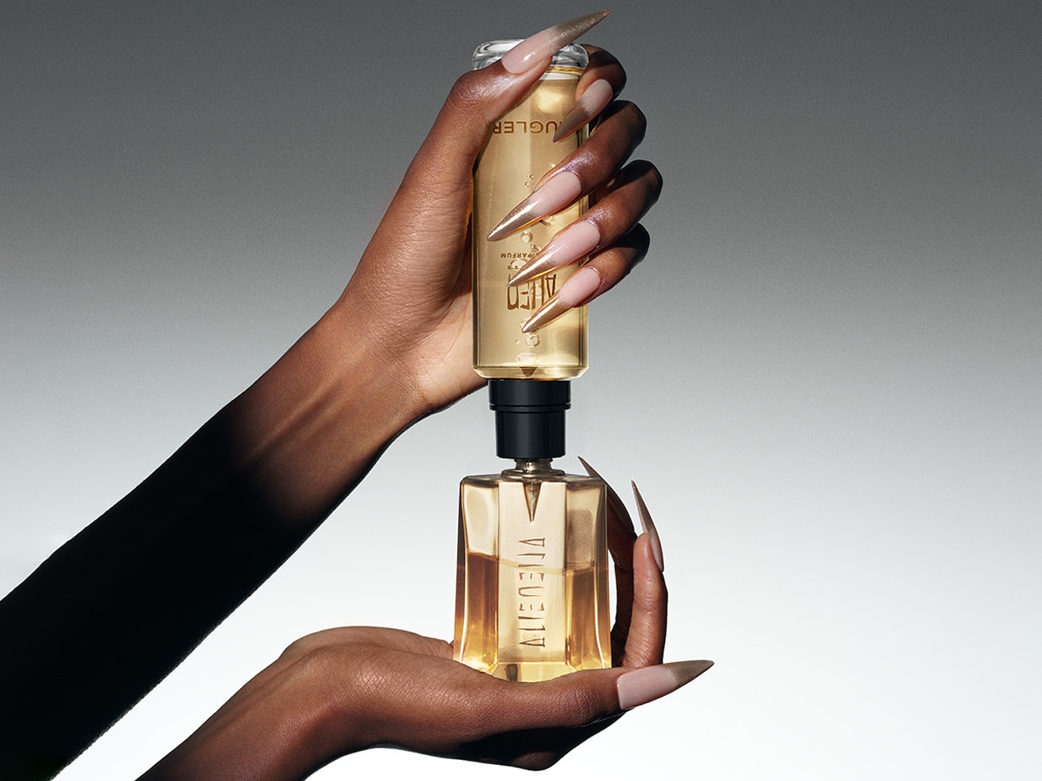 Les mains tiennent un flacon de parfum Alien d'une couleur dorée chaude, avec un flacon de recharge au-dessus, le tout sur un fond gris doux.