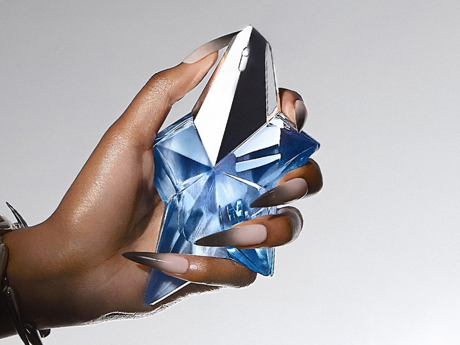 Main tenant un flacon de parfum bleu en forme d'étoile, un accessoire de poignet argenté ajoute une touche d'éclat.