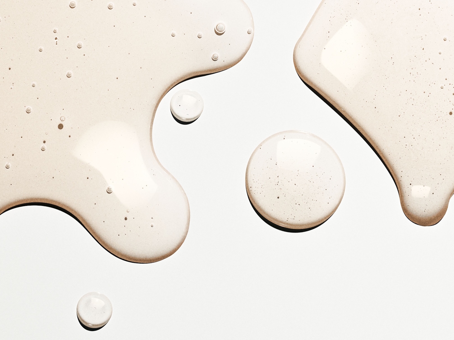 Nahaufnahme von transparenten, beigen Flüssigkeitstropfen und -pfützen auf einer glatten, hellen Oberfläche, die eine feuchtigkeitsspendende Textur und subtile Bläschen zeigt.