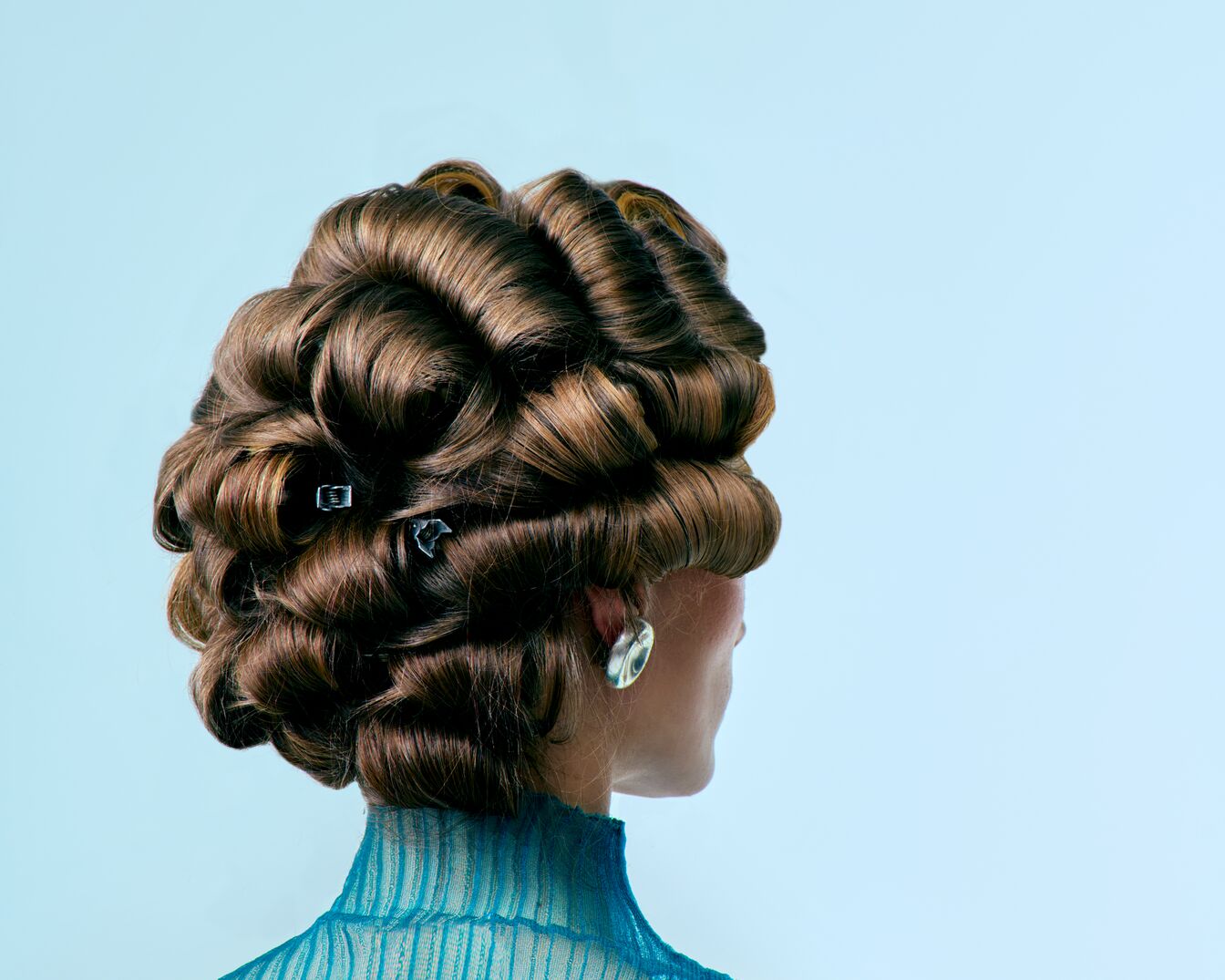 Vue arrière d'une coiffure élaborée avec des boucles brunes fixées par des épingles, complétée par une blouse turquoise et des boucles d'oreilles argentées.