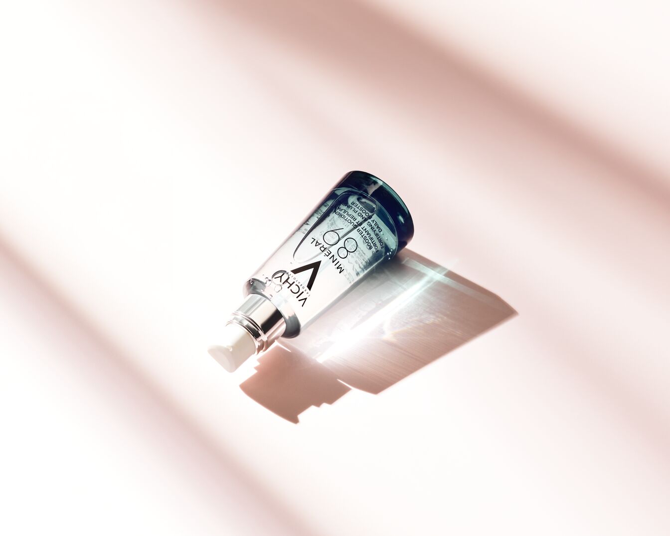 Das Vichy Minéral 89 Serum in einer durchsichtigen Glasflasche mit blauem Deckel und silbernem Spender liegt auf einer hellrosa Oberfläche und verspricht eine tägliche Stärkung und Aufpolsterung der Haut.