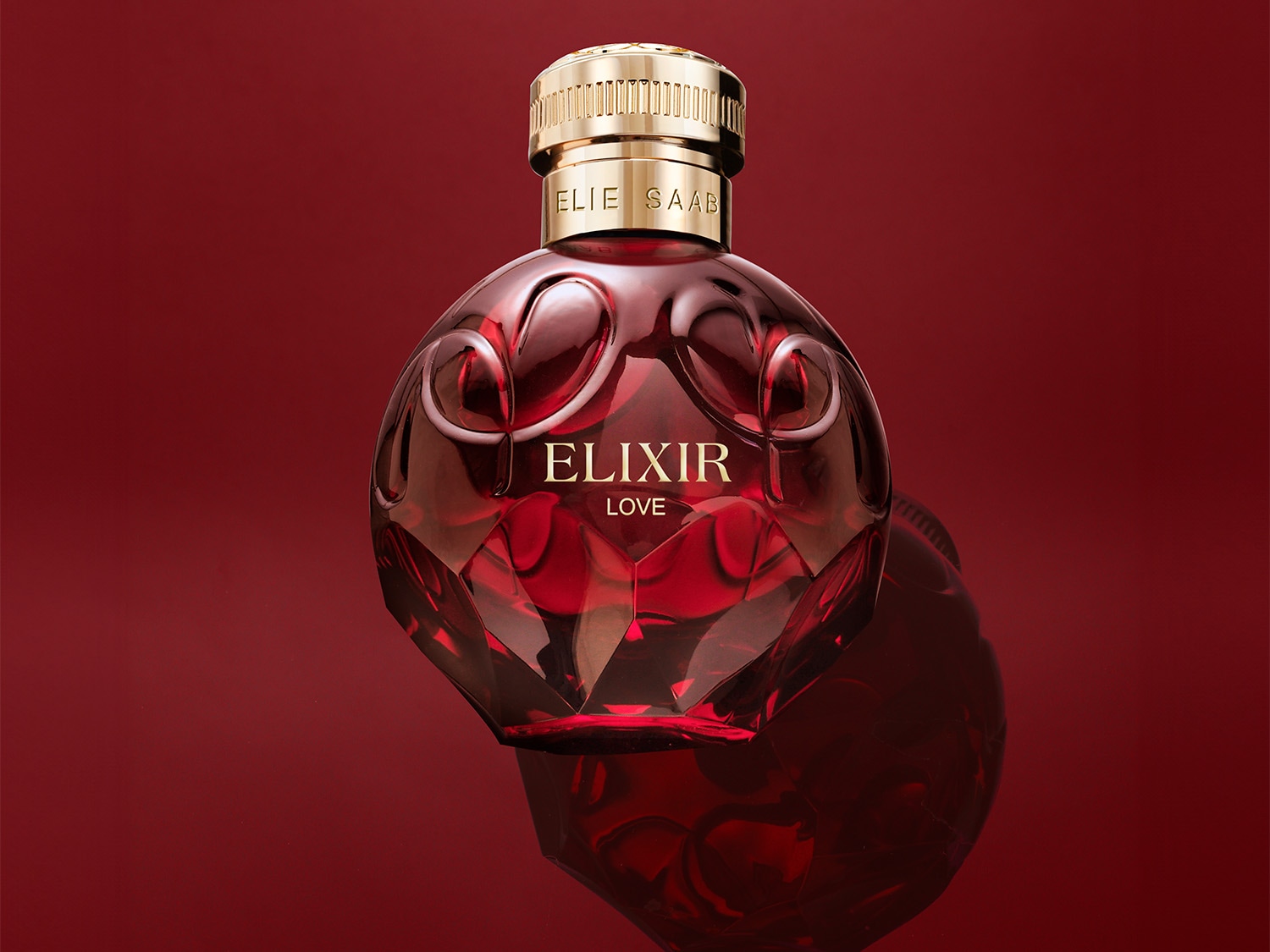 Gepresenteerd tegen een rode achtergrond, toont de ELIE SAAB Elixir Love parfumfles een bolvormig ontwerp met een gouden dop en de naam van het parfum in gouden letters.