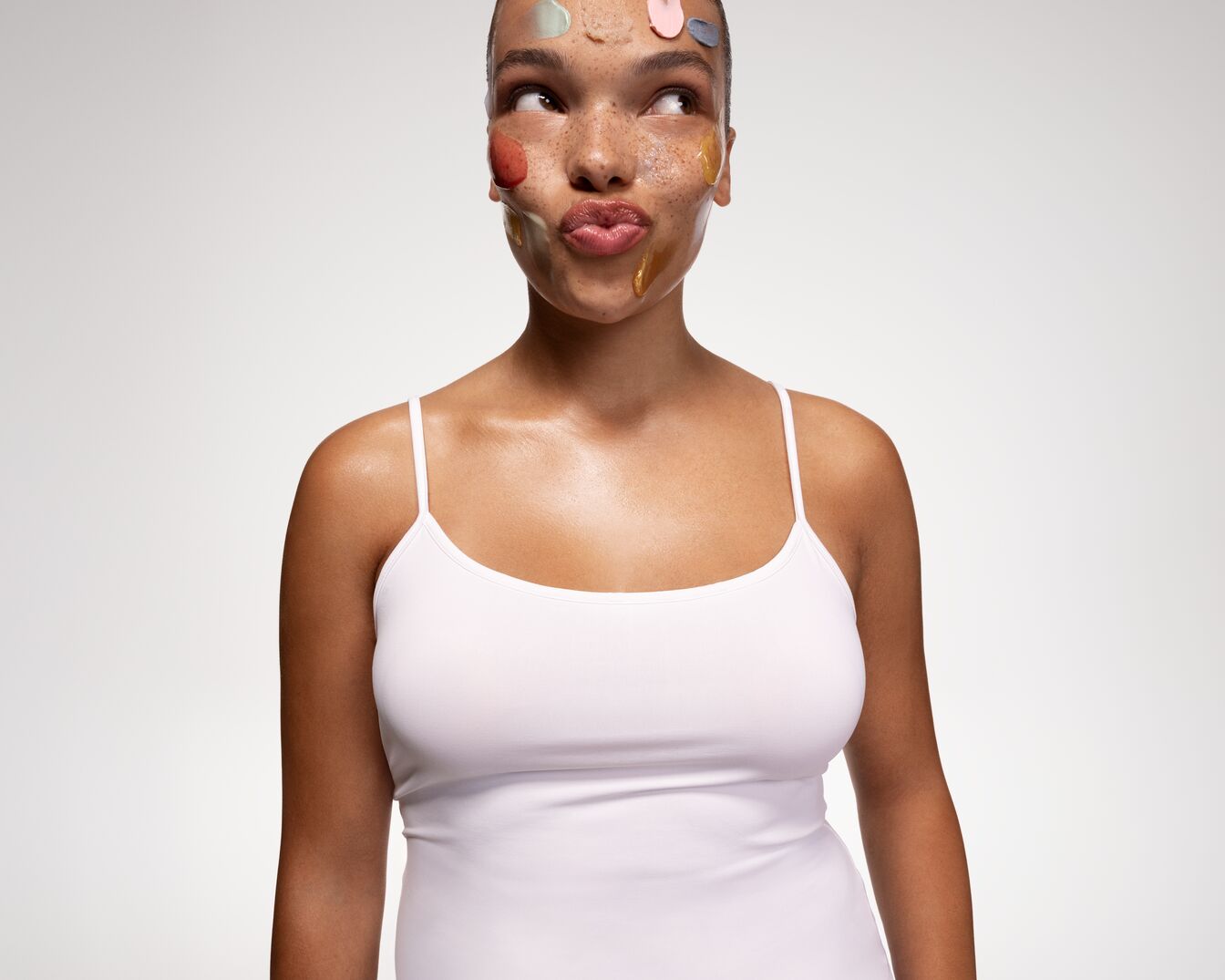 Eine Frau mit Sommersprossen und verschiedenen farbigen Masken auf ihrem Gesicht, die in einem weißen Top posiert und wegschaut.