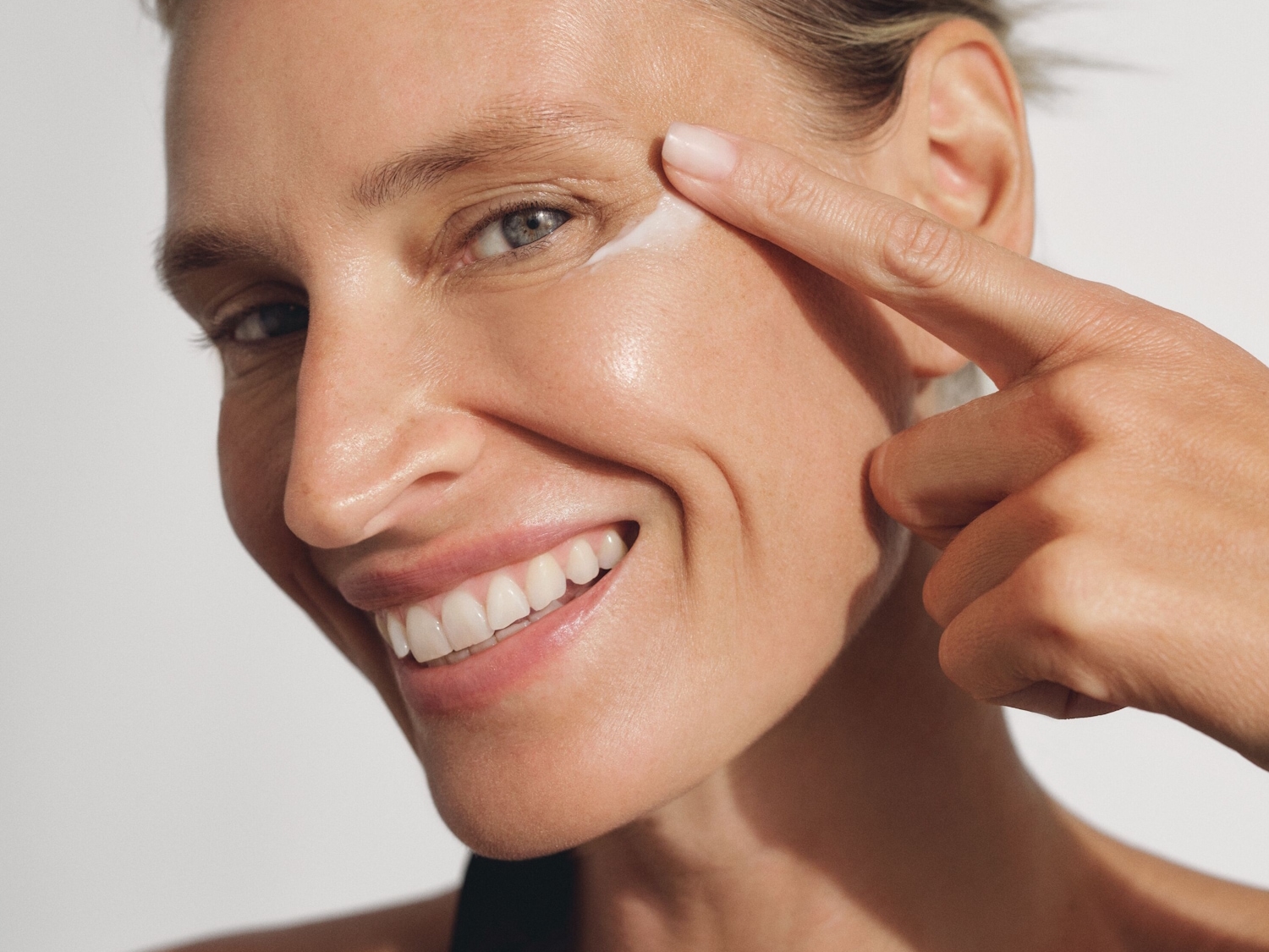 Primer plano de una mujer sonriente aplicando una crema blanca con el dedo índice en la zona de debajo del ojo.