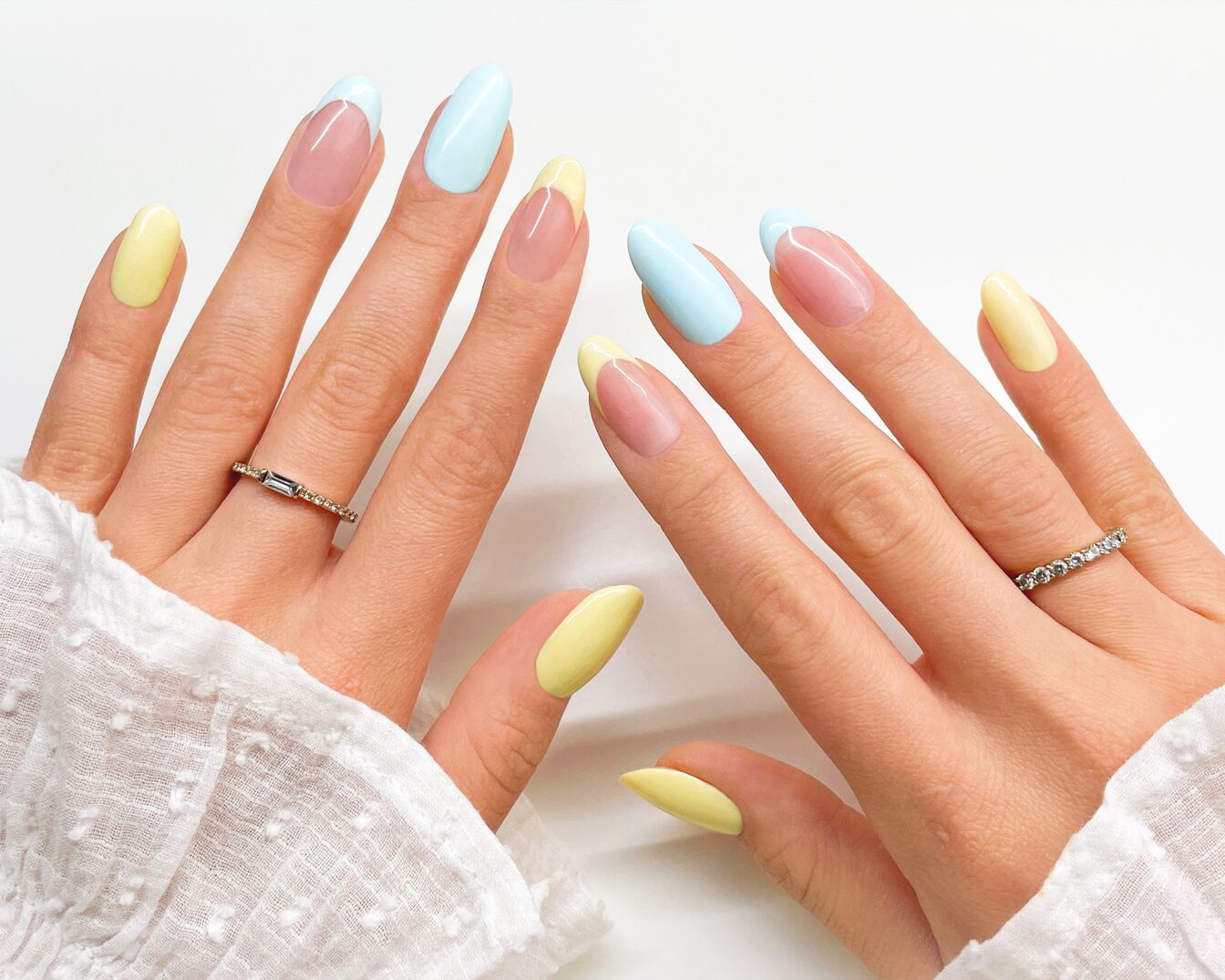 Las manos muestran uñas almendradas con esmalte pastel amarillo y azul, acentuadas con delicados anillos.