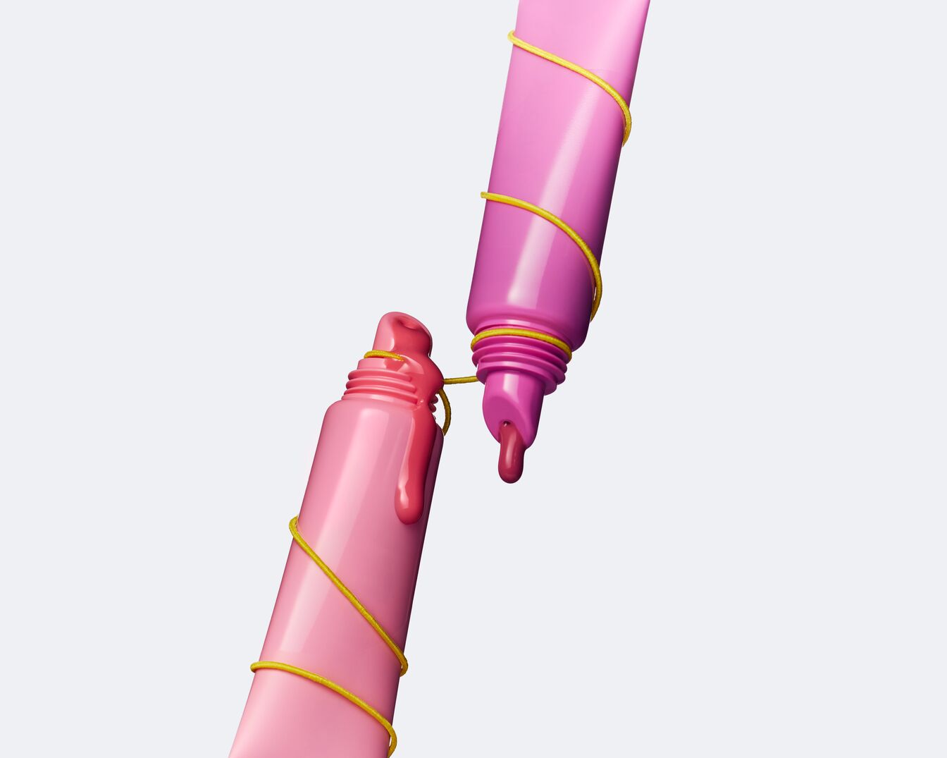 Dos tubos de brillo labial rosa, uno rosa claro y otro rosa oscuro, están conectados con una cuerda amarilla, mostrando una gota de producto.
