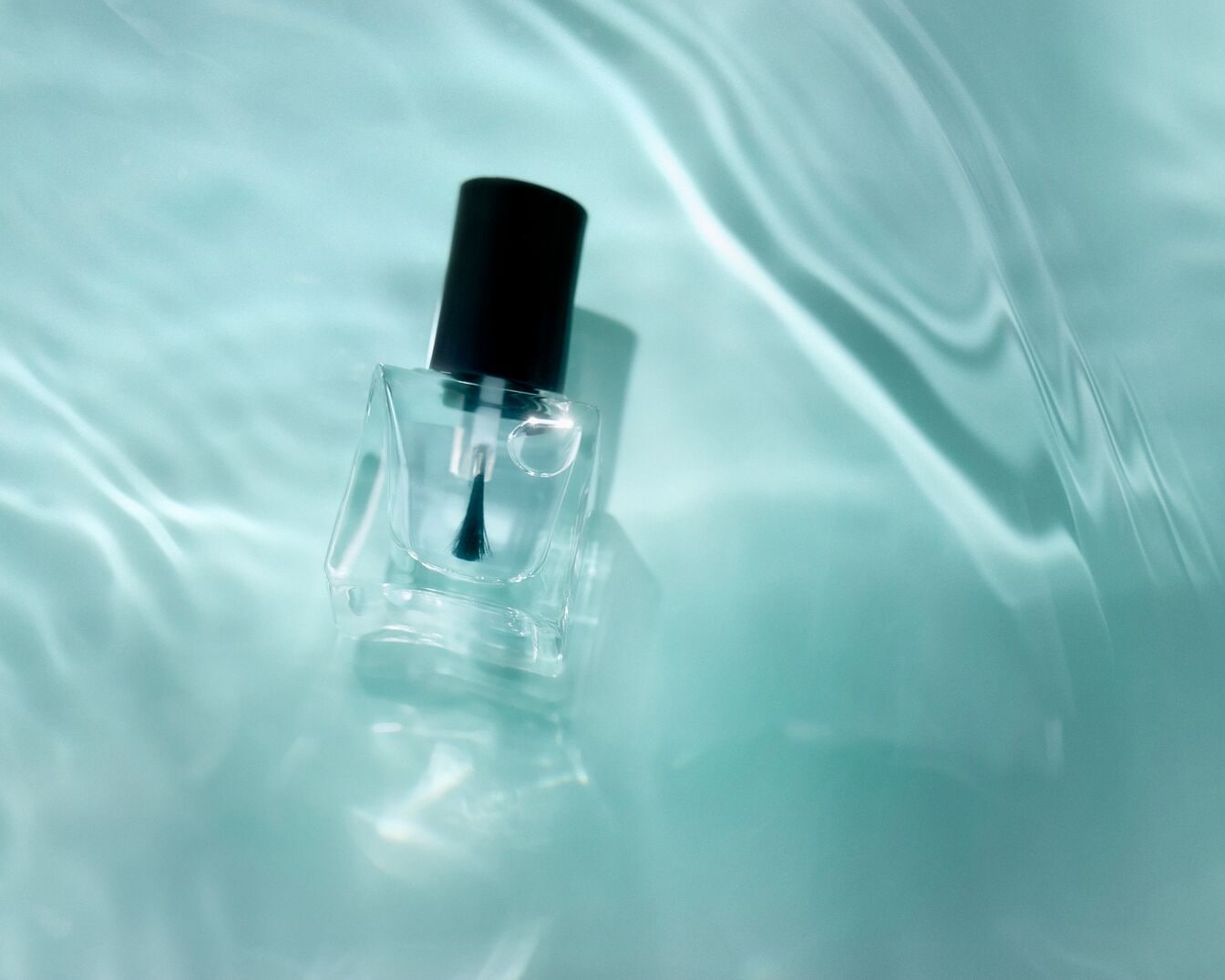 El esmalte de uñas transparente en un frasco de vidrio cuadrado con tapa negra, sobre un fondo azul claro ondulado, ofrece un brillo sutil para un acabado impecable.
