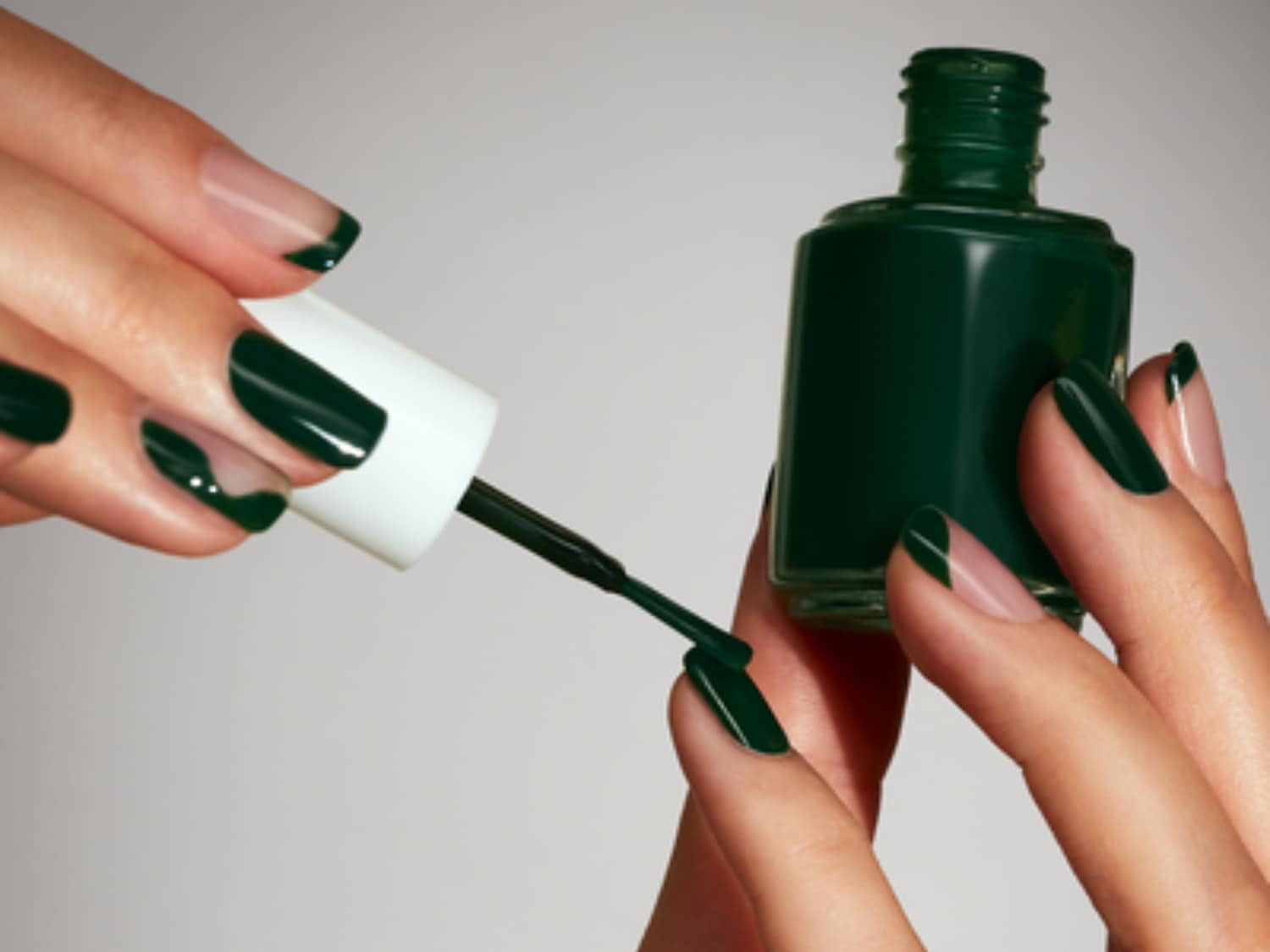 Aquí se muestra una botella de esmalte de uñas verde oscuro con una tapa blanca, mientras una mano aplica el esmalte en una uña, creando una manicura francesa verde.