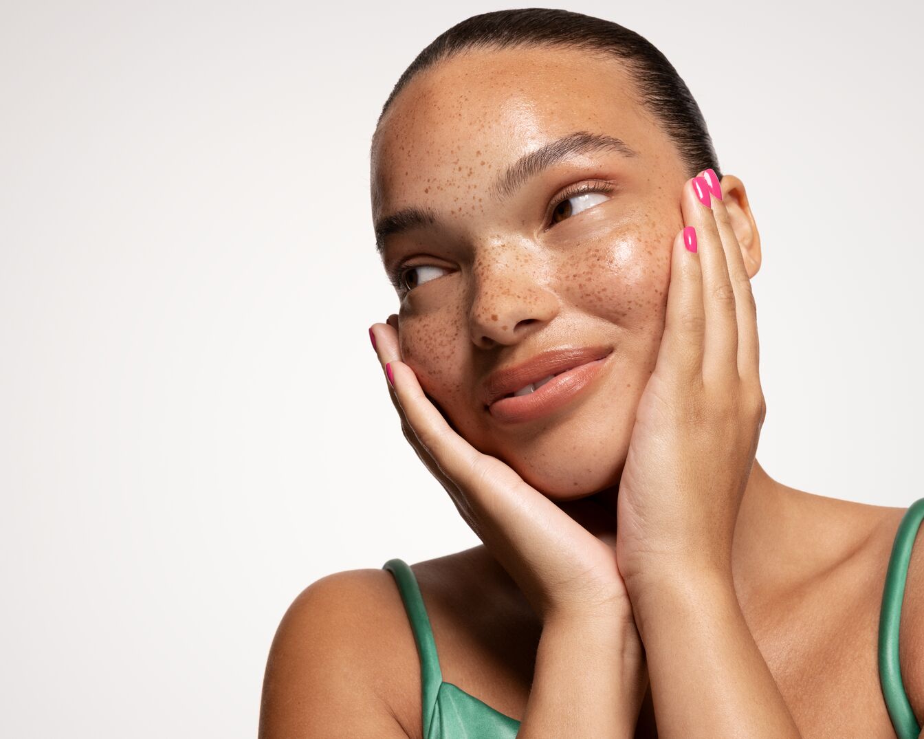 Una mujer de piel morena con pecas y uñas rosas sostiene su rostro con ambas manos, luciendo una piel brillante y un labial nude.