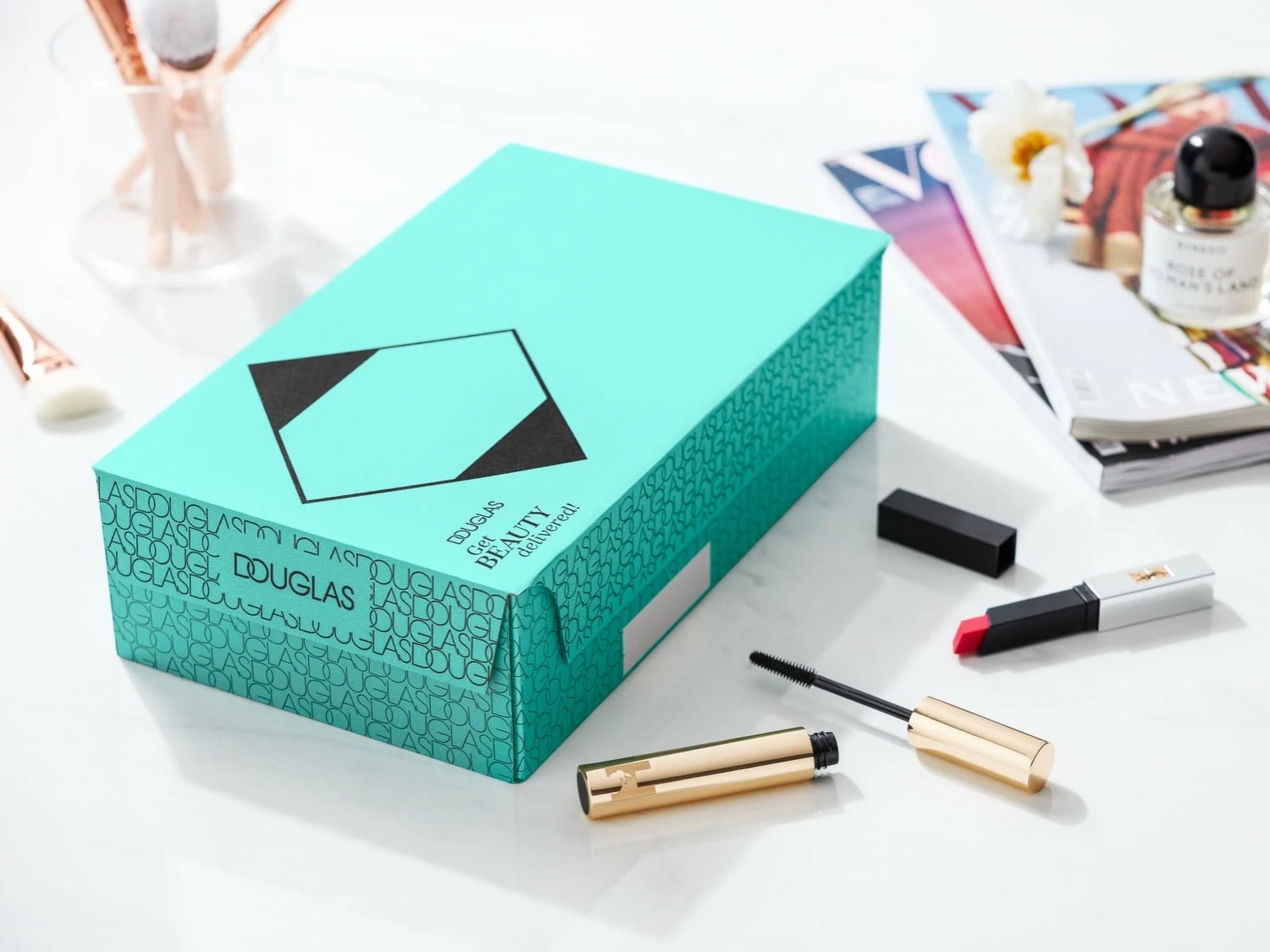 Scatola regalo turchese di Douglas con mascara dorato aperto, rossetto e pennelli da trucco sullo sfondo.