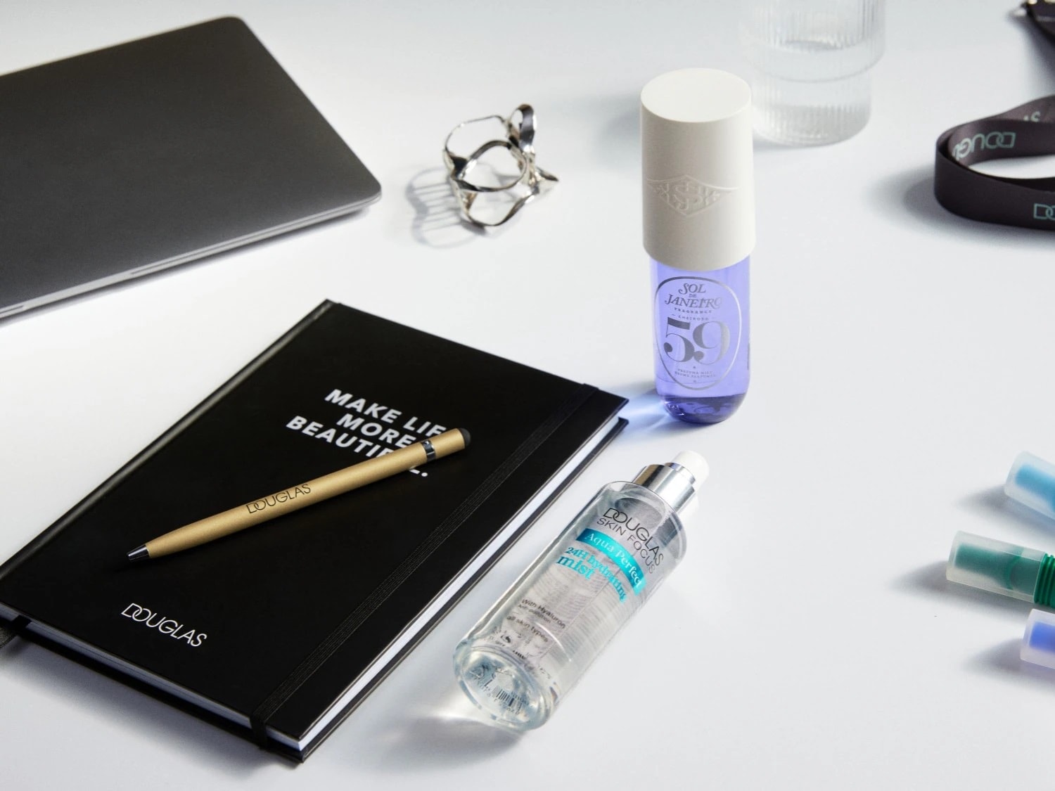 Un'agenda nera con la scritta 'MAKE LIFE MORE BEAUTIFUL' e una penna dorata Douglas, accanto a prodotti di bellezza come lo spray profumato Sol de Janeiro 59 e la brume idratante Aqua Perfect di Douglas.