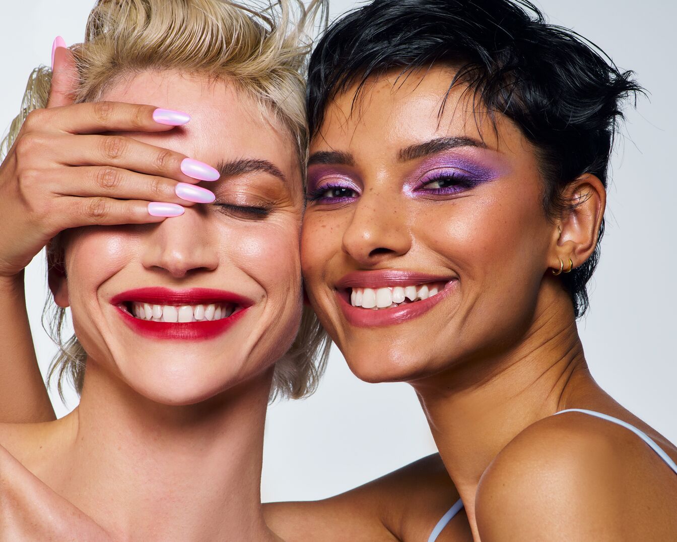 Zwei strahlende Frauen mit lebendigem Make-up: Die eine mit rotem Lippenstift und schimmernden Nägeln, die andere mit auffälligem lila Lidschatten.