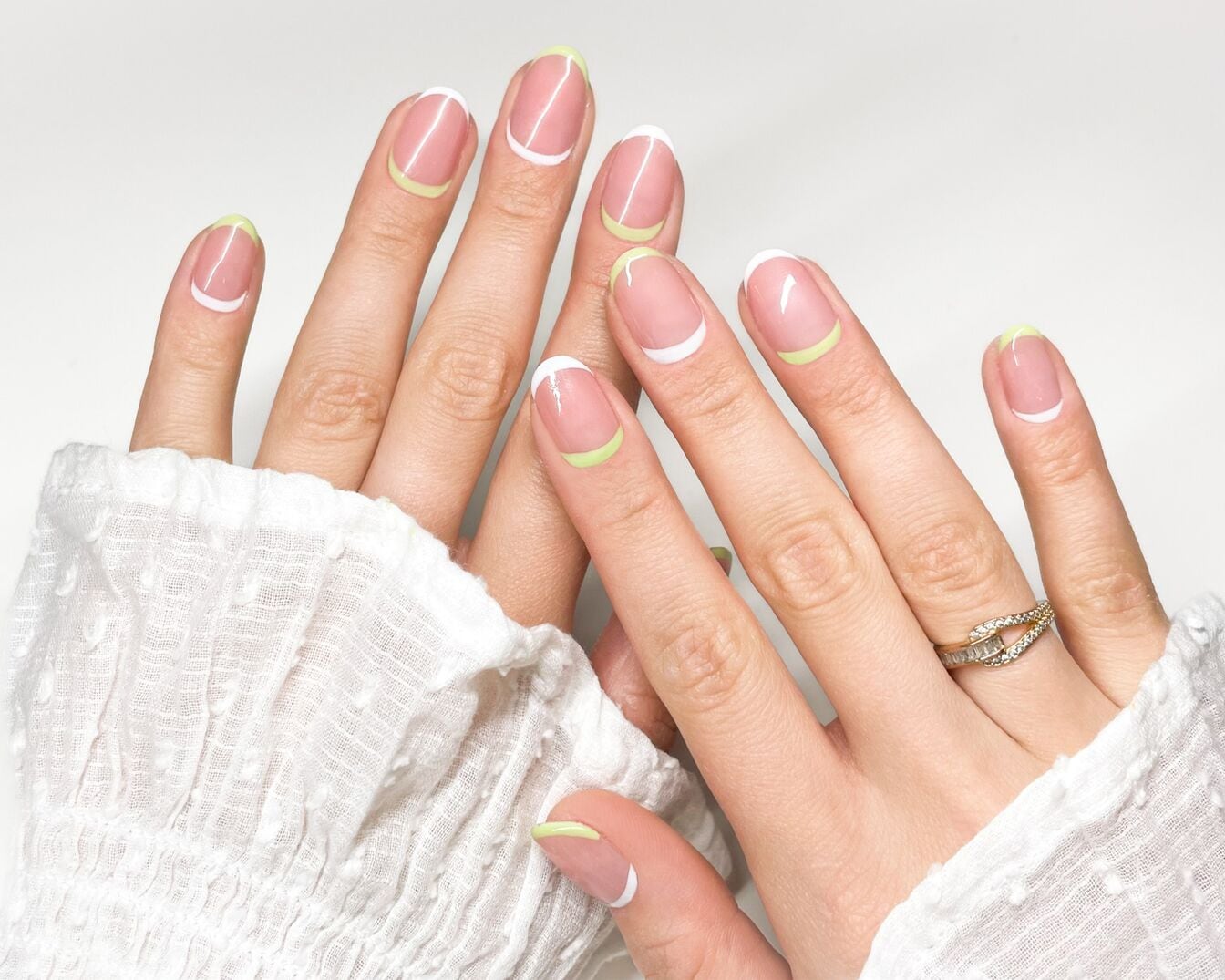 Nahaufnahme von zwei Händen mit mandelförmigen Nägeln, die mit einem weißen und hellgrünen French-Look verziert sind, wobei die rechte Hand einen goldenen Ring trägt.