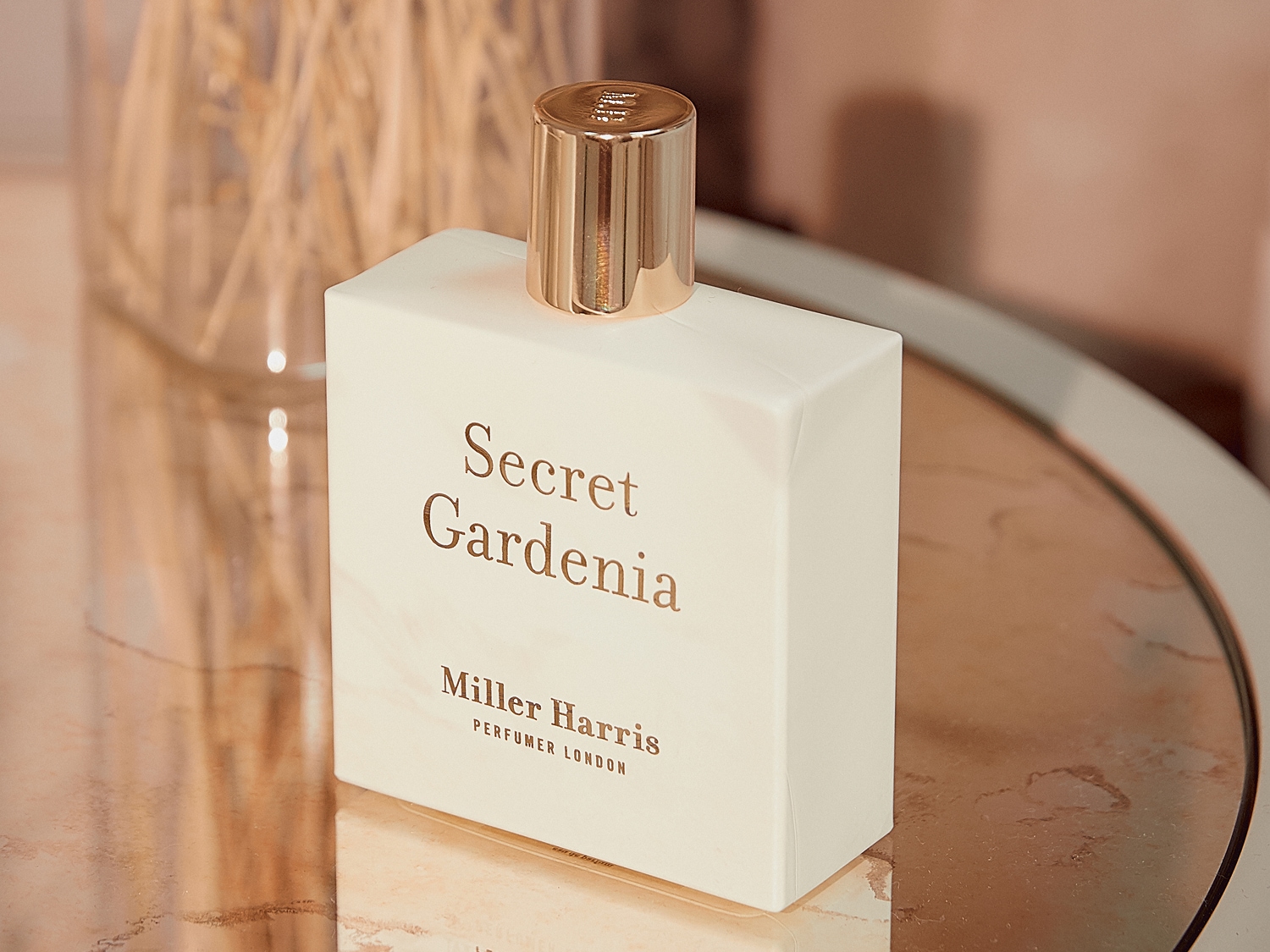 Biały flakon perfum Secret Gardenia marki Miller Harris z błyszczącą, złotą nakrętką, stojący na błyszczącej powierzchni.
