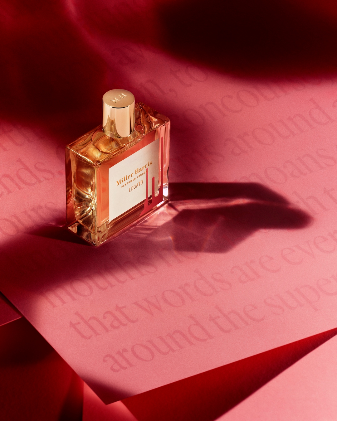 Butelka perfum Miller Harris Legato o bursztynowym zabarwieniu z złotą nakrętką stoi na różowym tle z napisem, tworząc elegancką kompozycję.