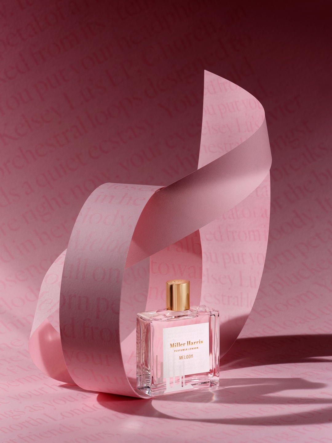 Różowy flakon perfum Miller Harris Melody z złotą nakrętką, otoczony wstążką na tle z tekstem, tworząc delikatną i elegancką kompozycję.