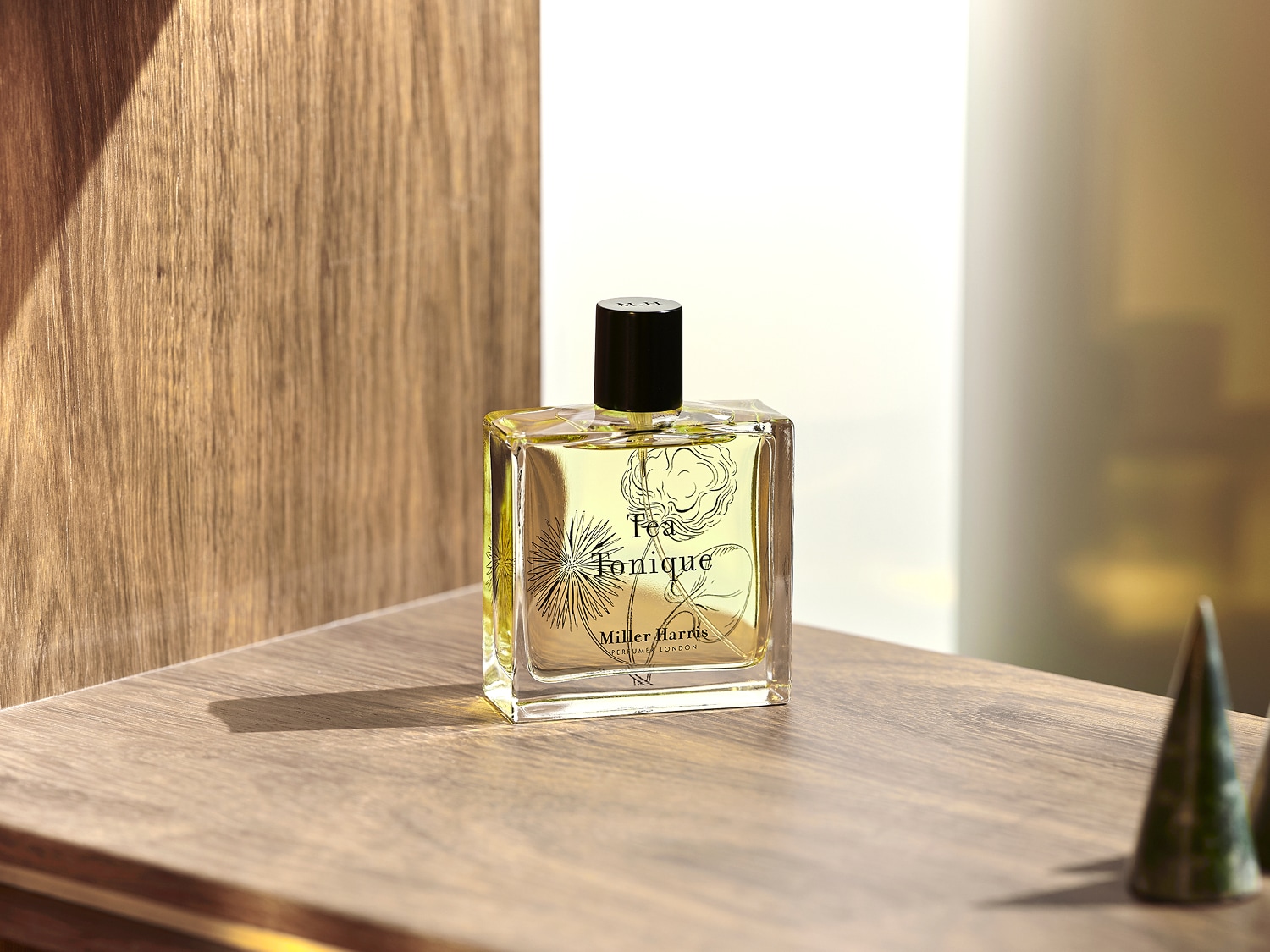 Przezroczysty flakon perfum Tea Tonique Miller Harris z czarnym korkiem stoi na drewnianej powierzchni, ukazując delikatny, kwiatowy wzór na etykiecie.