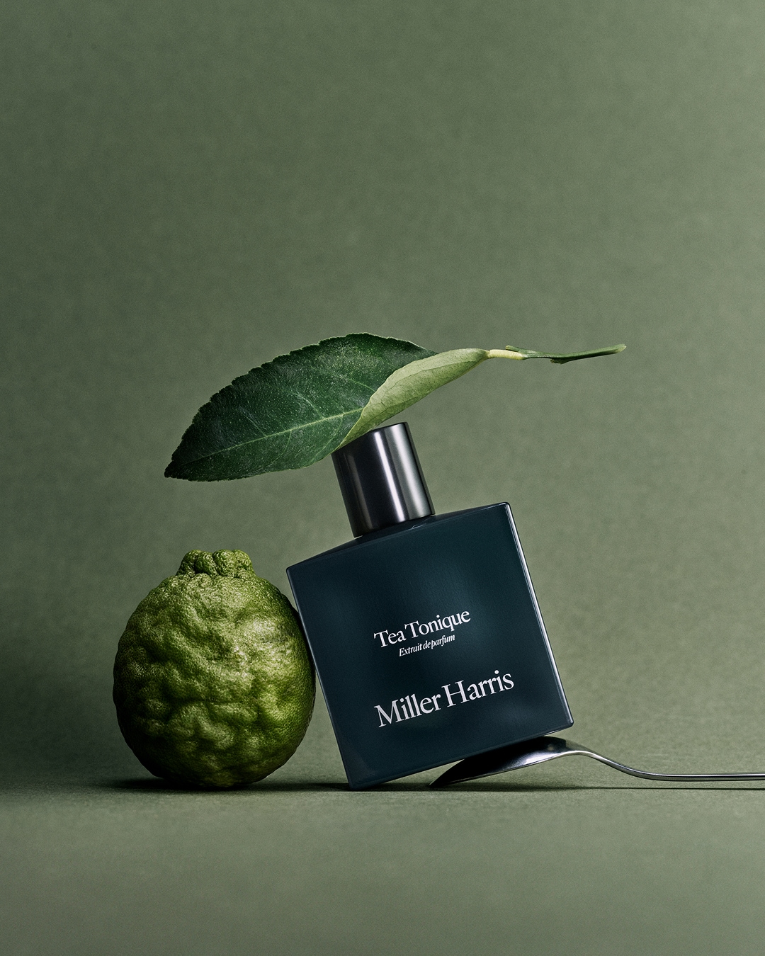 Butelka perfum Tea Tonique Extrait de parfum marki Miller Harris stoi obok zielonej limonki i liścia na zielonym tle.