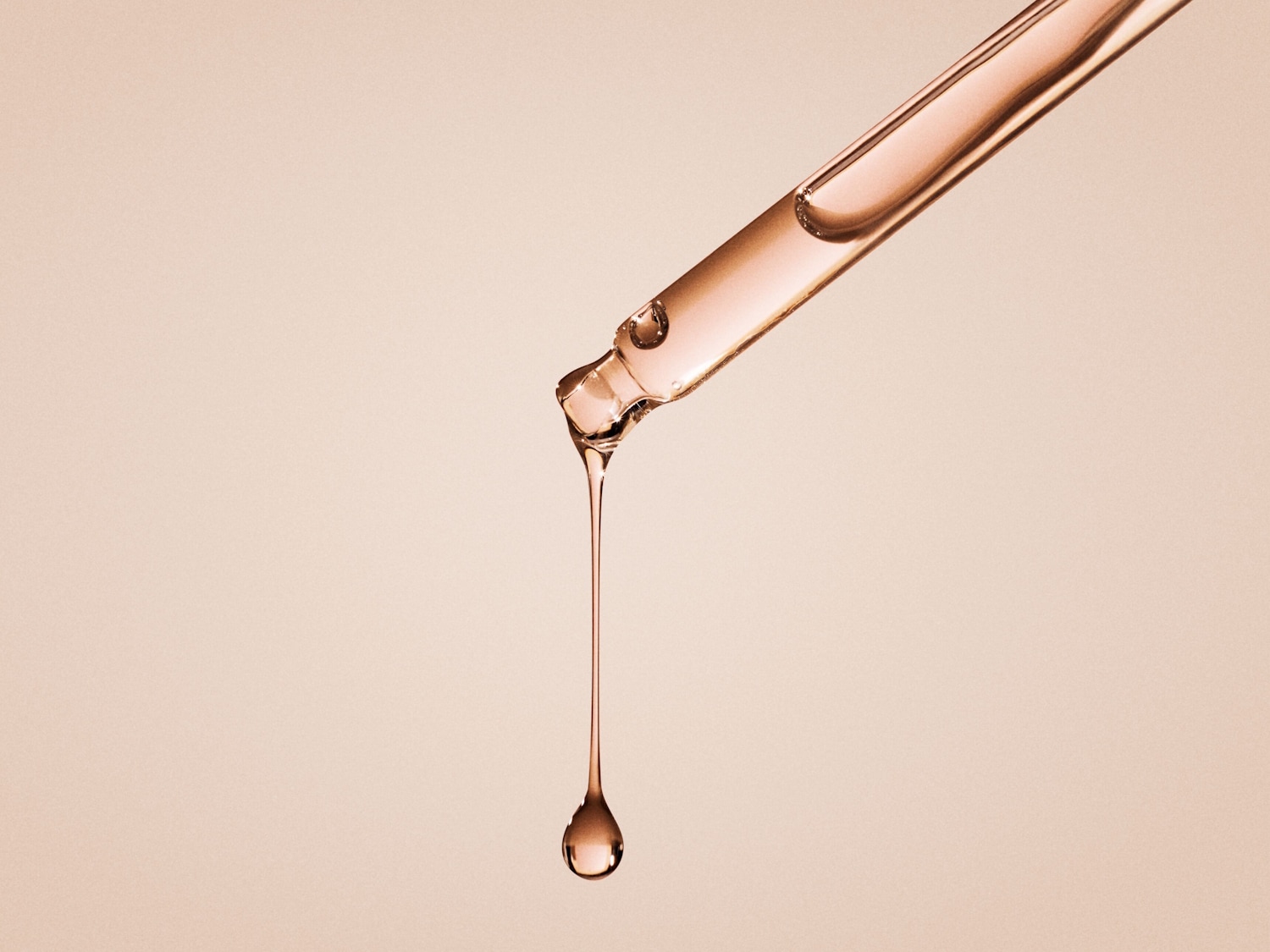 Nahaufnahme einer Pipette, aus der ein Tropfen eines klaren, rosafarbenen Serums austritt, das die Essenz von Feuchtigkeit und Verjüngung für strahlende Haut verspricht.