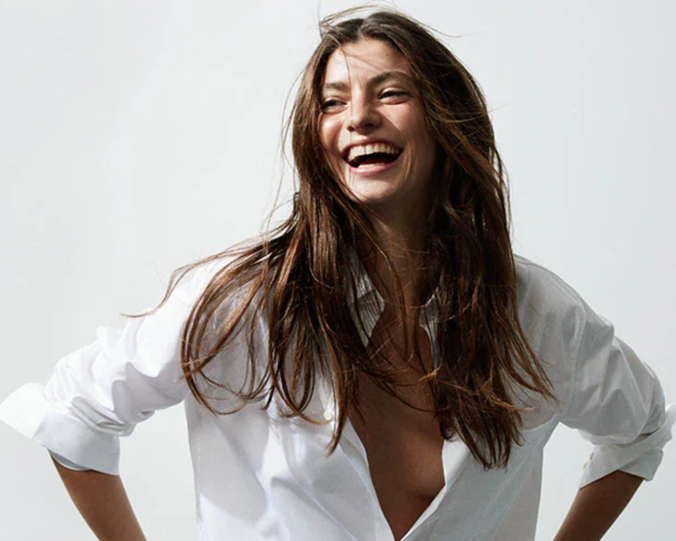 Una mujer sonriente con cabello castaño suelto y una camisa blanca desabrochada irradia alegría.