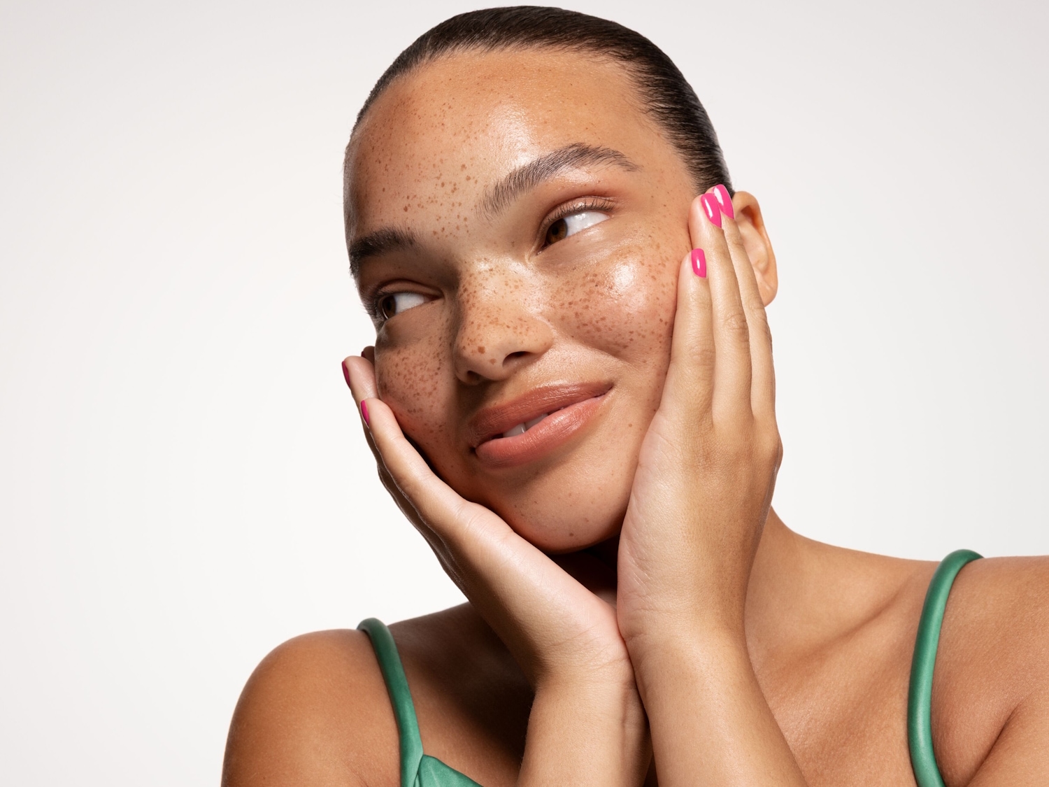 Una mujer sonriente con pecas y uñas rosas sostiene su rostro con las manos, mostrando una piel radiante y un aspecto saludable.