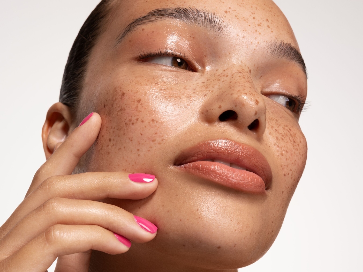 Primer plano del rostro de una mujer con pecas, piel brillante y labios color melocotón, tocándose suavemente la mejilla con la mano de uñas rosas.
