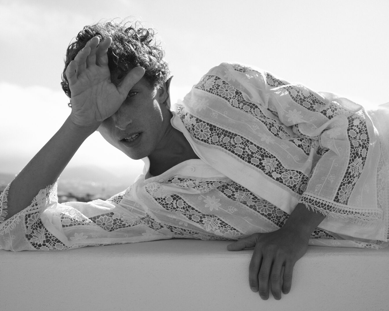En una foto en blanco y negro, un hombre con cabello rizado y una camisa de encaje blanca está acostado, con la mano en la frente, creando una imagen elegante y relajada.
