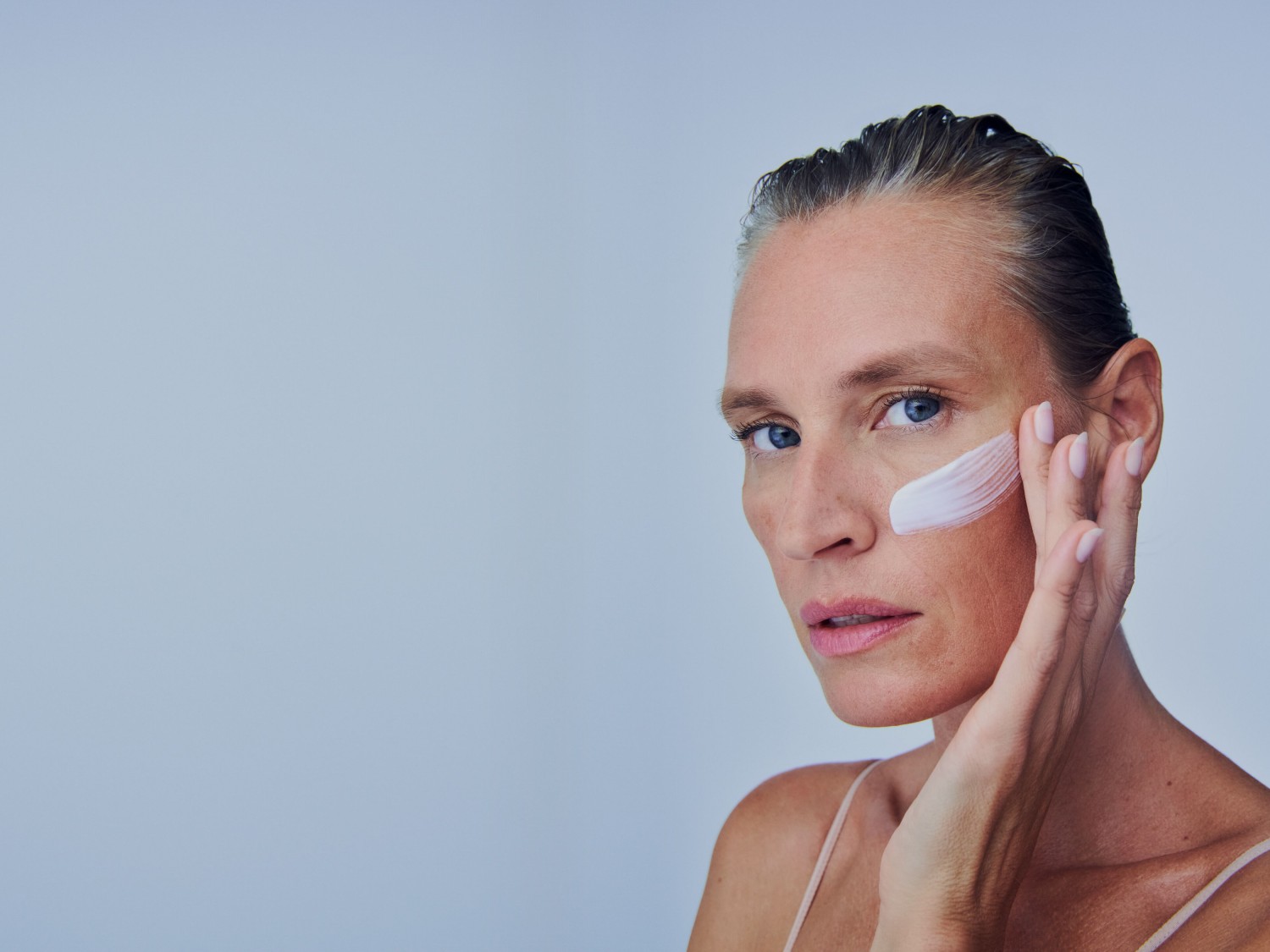 Eine Frau mit hellblauen Augen trägt eine weiße Creme auf ihre Wange auf und schaut in die Kamera.