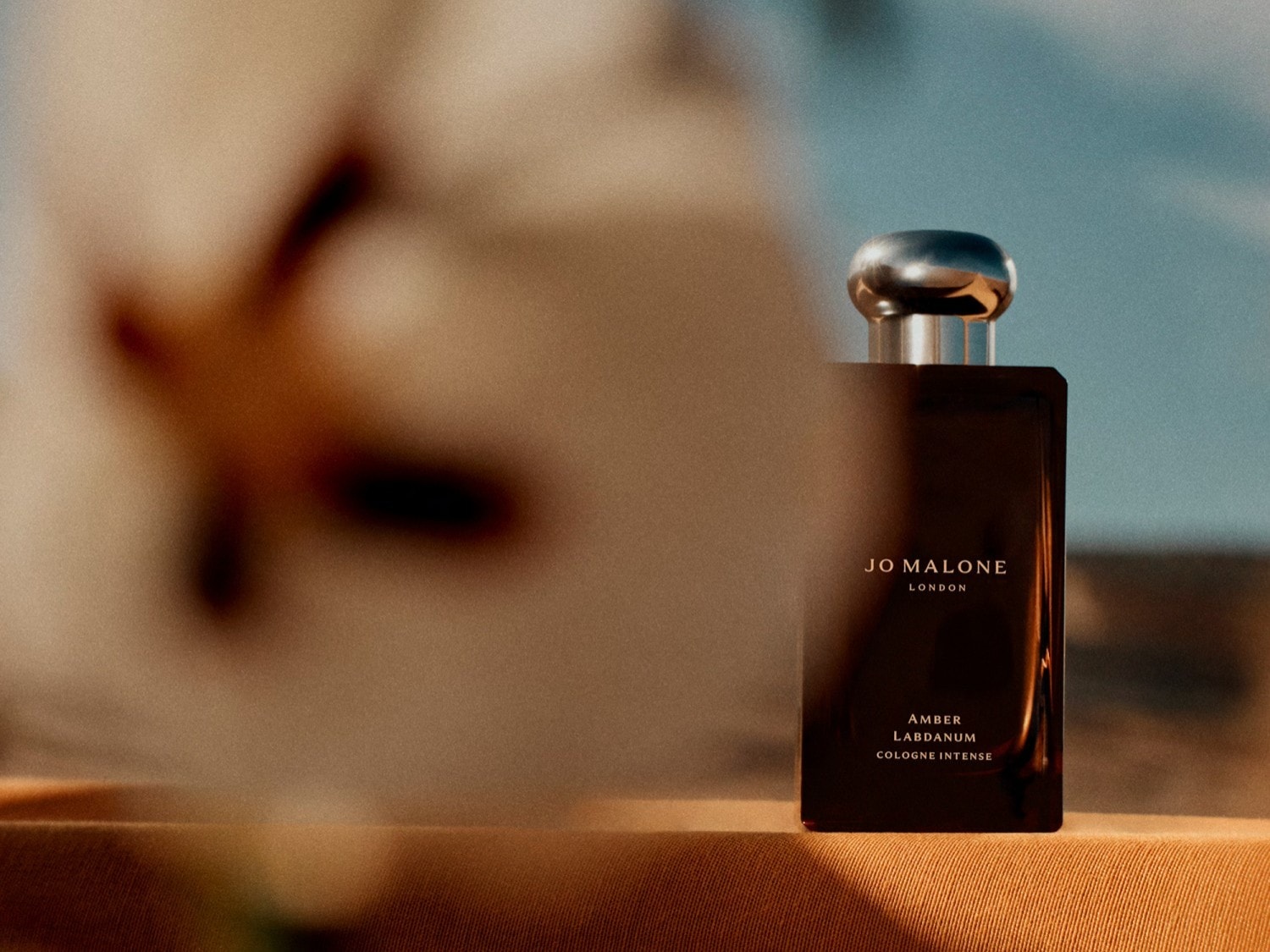 Butelka Jo Malone London Amber Labdanum Cologne Intense w bursztynowym odcieniu, z metalową nakrętką, stoi na tle nieba i abstrakcyjnego kształtu kwiatu.