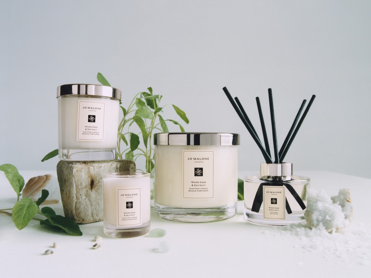 Kolekcja Jo Malone London Wood Sage & Sea Salt obejmuje świece zapachowe w szklanych słojach z srebrnymi pokrywkami i dyfuzor zapachowy z czarnymi patyczkami.