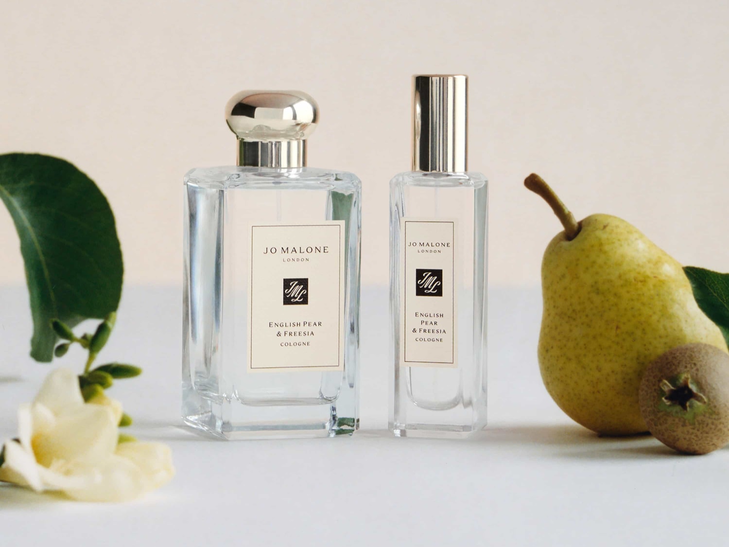 Dwa flakony perfum Jo Malone English Pear & Freesia Cologne stoją obok siebie, otoczone świeżą gruszką, kwiatami frezji i zielonymi liśćmi, tworząc elegancką kompozycję.