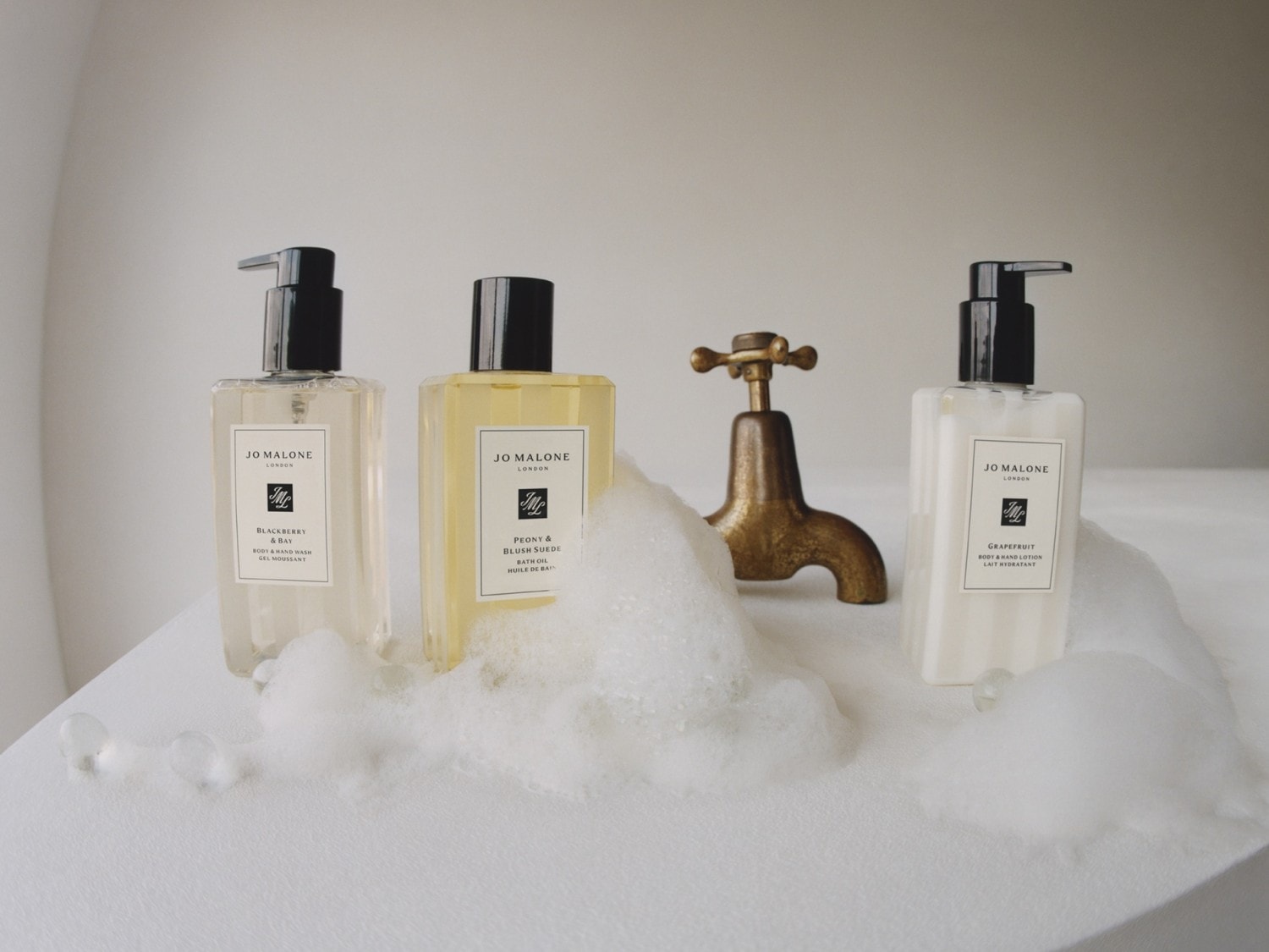 Trzy butelki produktów Jo Malone: żel do mycia ciała i rąk Blackberry & Bay, olejek do kąpieli Peony & Blush Suede oraz balsam do ciała i rąk Grapefruit, ustawione na białej powierzchni z pianą i złotym kranem.