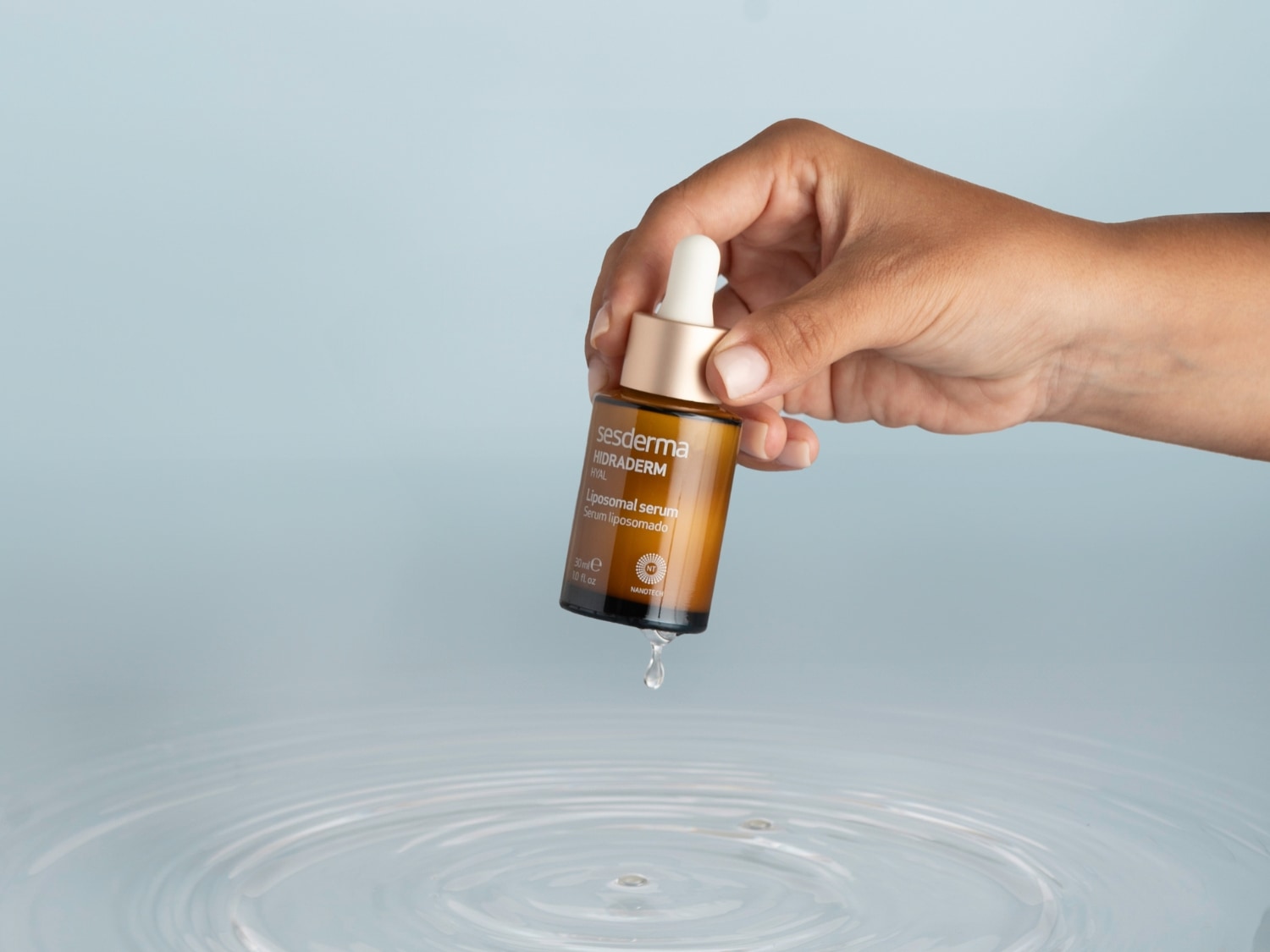 Dłoń trzyma butelkę serum Sesderma Hidraderm Hyal Liposomal Serum, z której kapie kropla, co sugeruje intensywne nawilżenie.