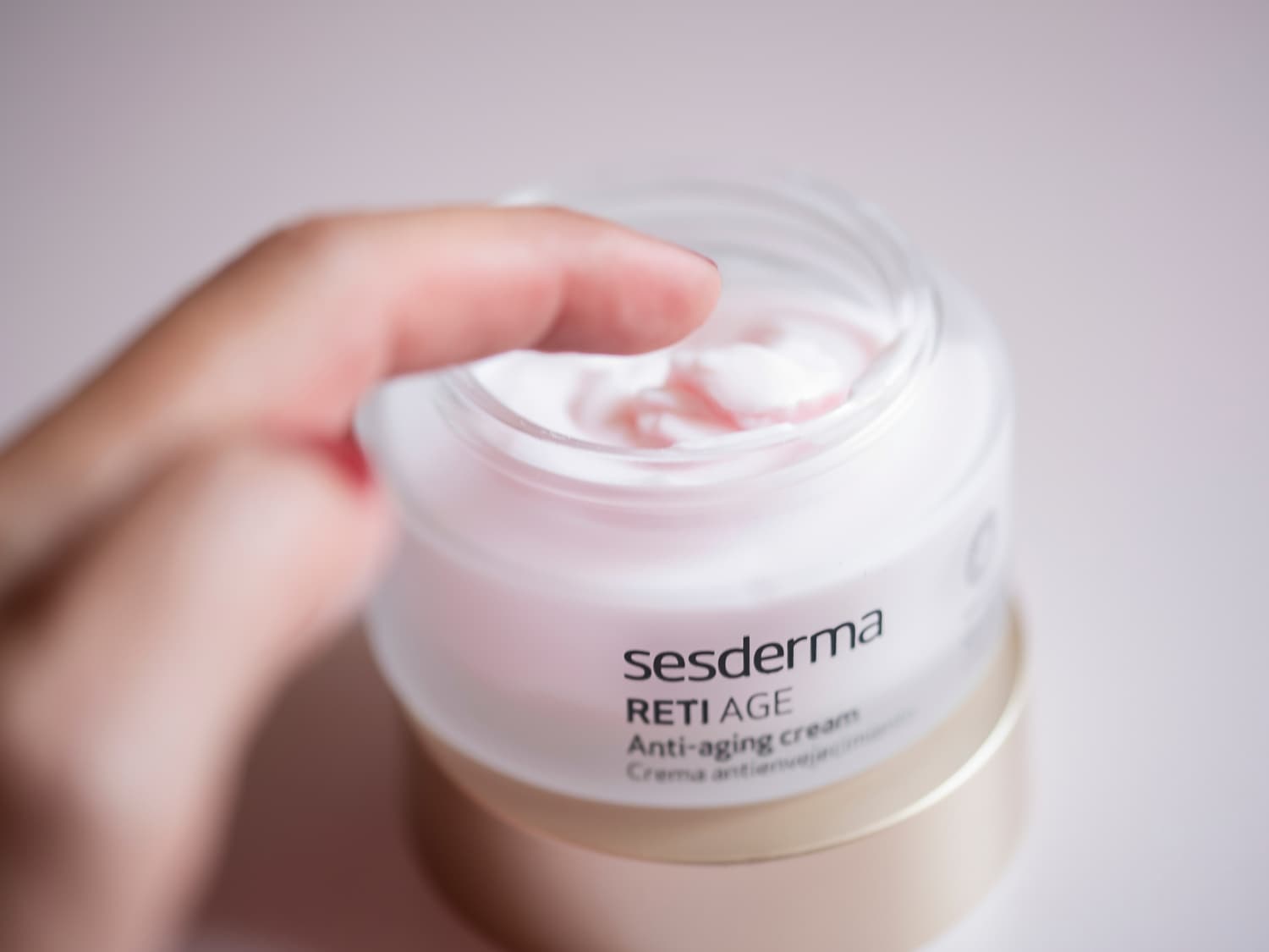Palec zanurza się w słoiczku z kremem Sesderma Reti Age Anti-aging o lekkiej, różowej konsystencji.