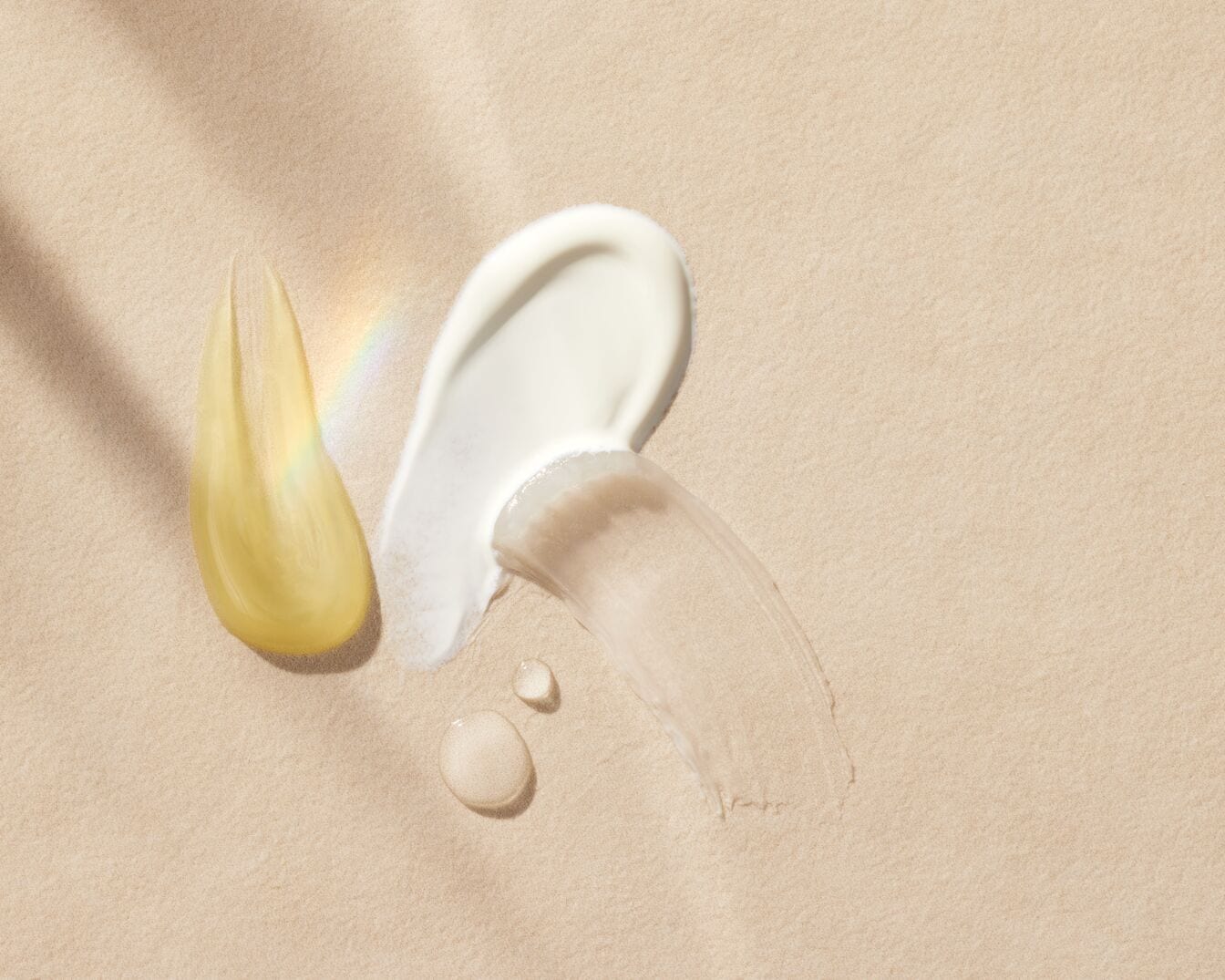 Na beżowym tle widoczne są próbki kosmetyków: złoty olejek, biały krem i beżowy fluid, które prezentują różne tekstury i odcienie produktów pielęgnacyjnych.