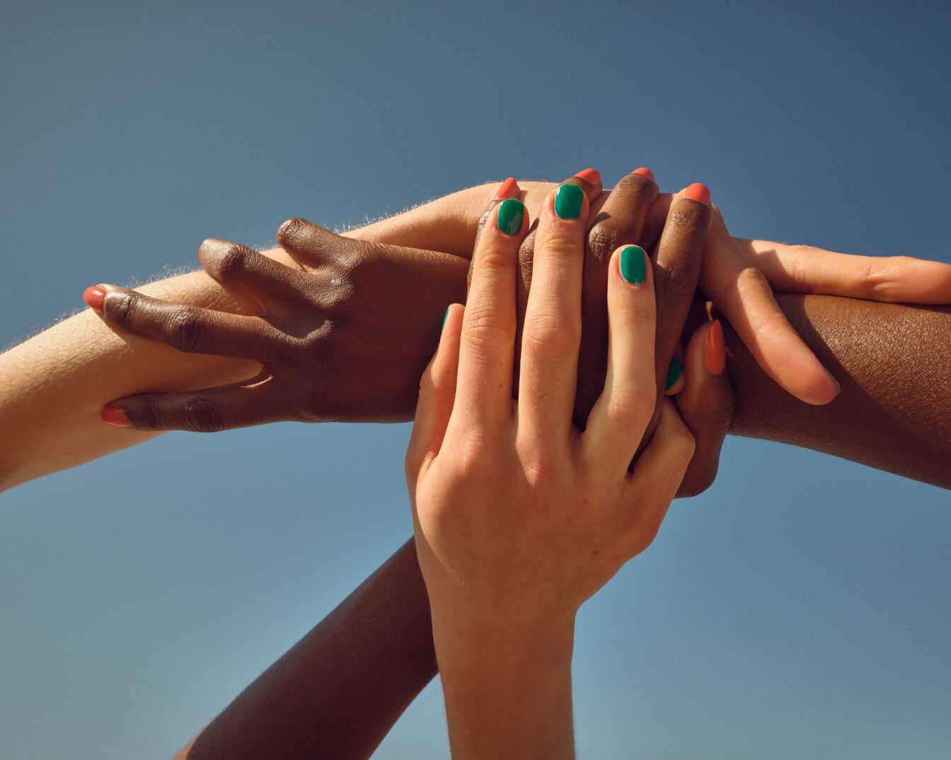 Na tle błękitnego nieba splecione dłonie o różnych odcieniach skóry, z paznokciami pomalowanymi na zielono i pomarańczowo, symbolizują jedność i różnorodność.