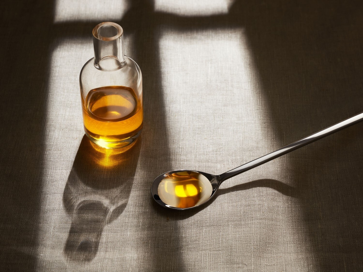 Klares Glasfläschchen mit goldfarbenem Öl neben einem Löffel, der ebenfalls mit Öl gefüllt ist, auf einem strukturierten Untergrund.